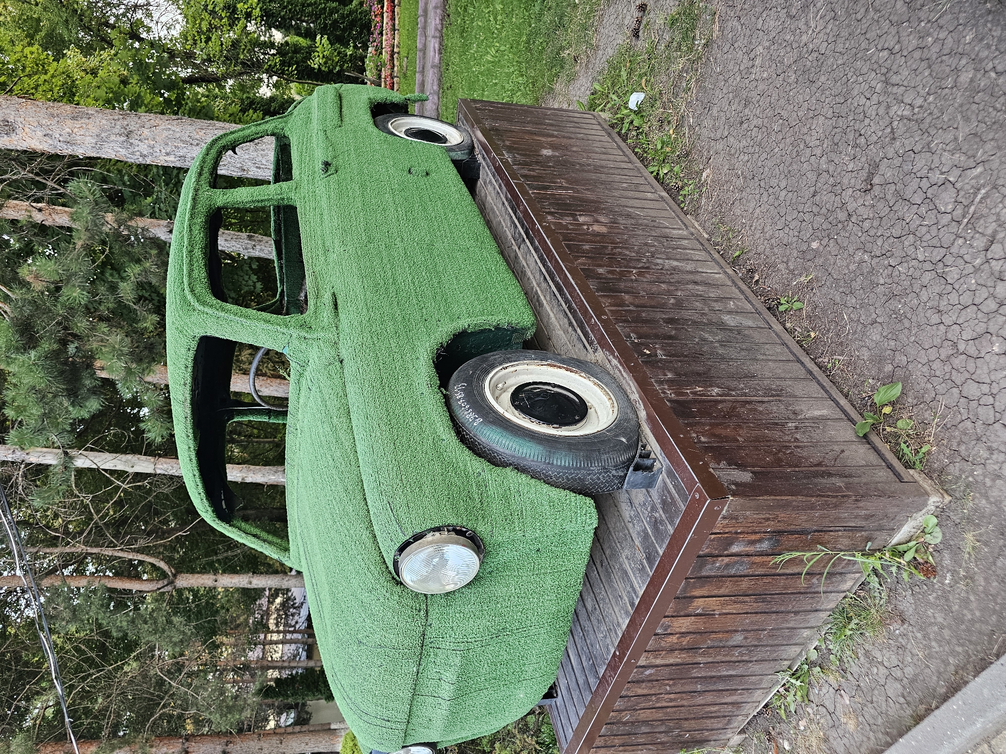 A vintage car, entirely covered in artificial green turf, is displayed on a wooden platform outdoors. The car appears to be a small, two-door hatchback. The platform is constructed of dark brown wooden planks with metal trim. The car is positioned in what appears to be a park or outdoor recreational area, with trees and grass in the background. The ground around the platform is a mixture of cracked earth and sparse grass. There are some pine cones visible on the ground. The scene is lit by natural daylight, suggesting it is daytime. The specific location is identified as Cherkessk, Russia. There are no people visible in the image.
