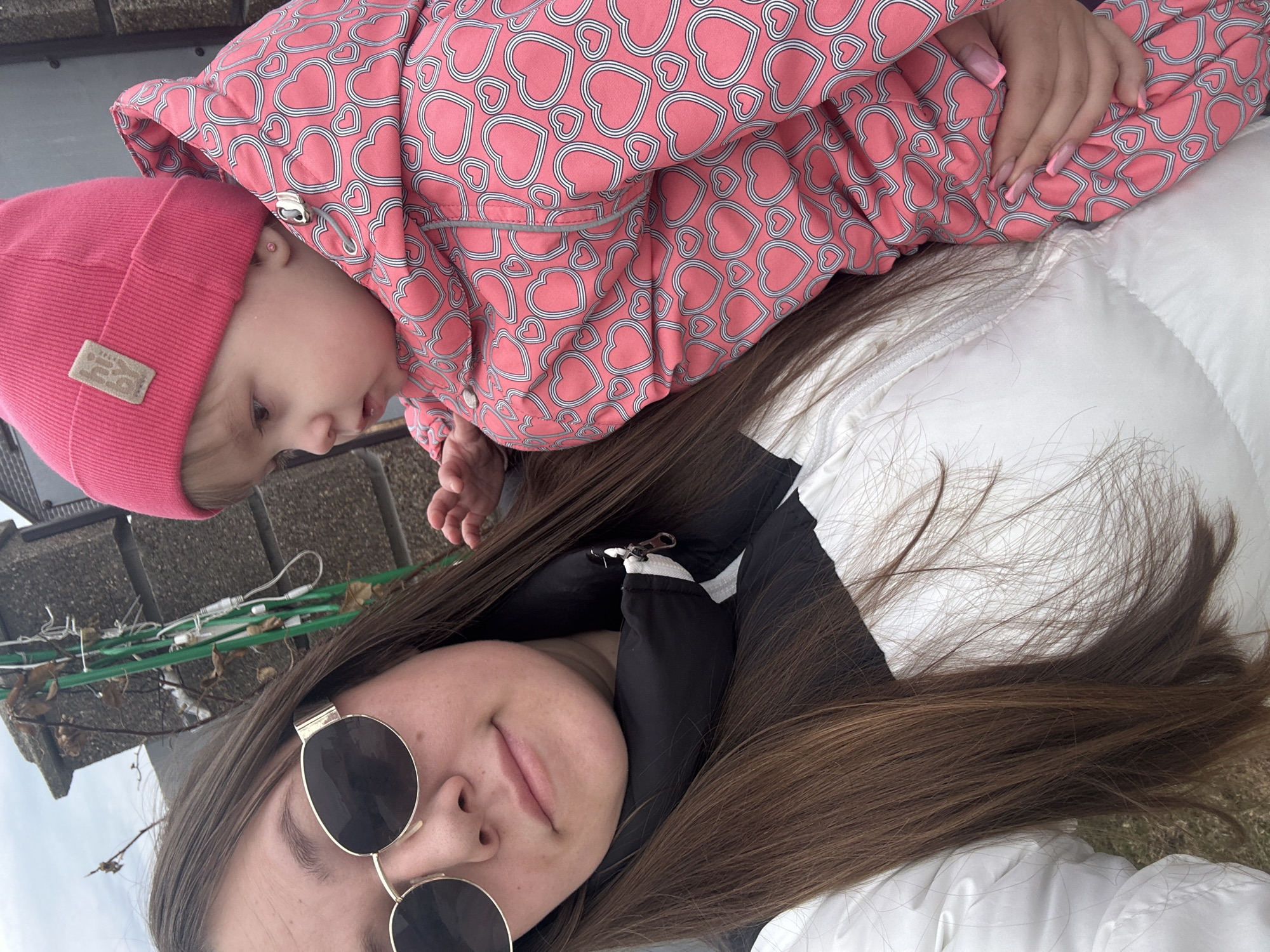A selfie shows an adult woman with long, brown hair, wearing round sunglasses, and a white jacket with black accents, lying on her back with a baby on top of her. The baby is wearing a pink hat and a pink jacket with a white heart pattern. The woman is looking at the camera with a slight smile. The baby is looking to the left, slightly downward. The background shows a brick wall and some string lights. The location is Ольховка, Russia.