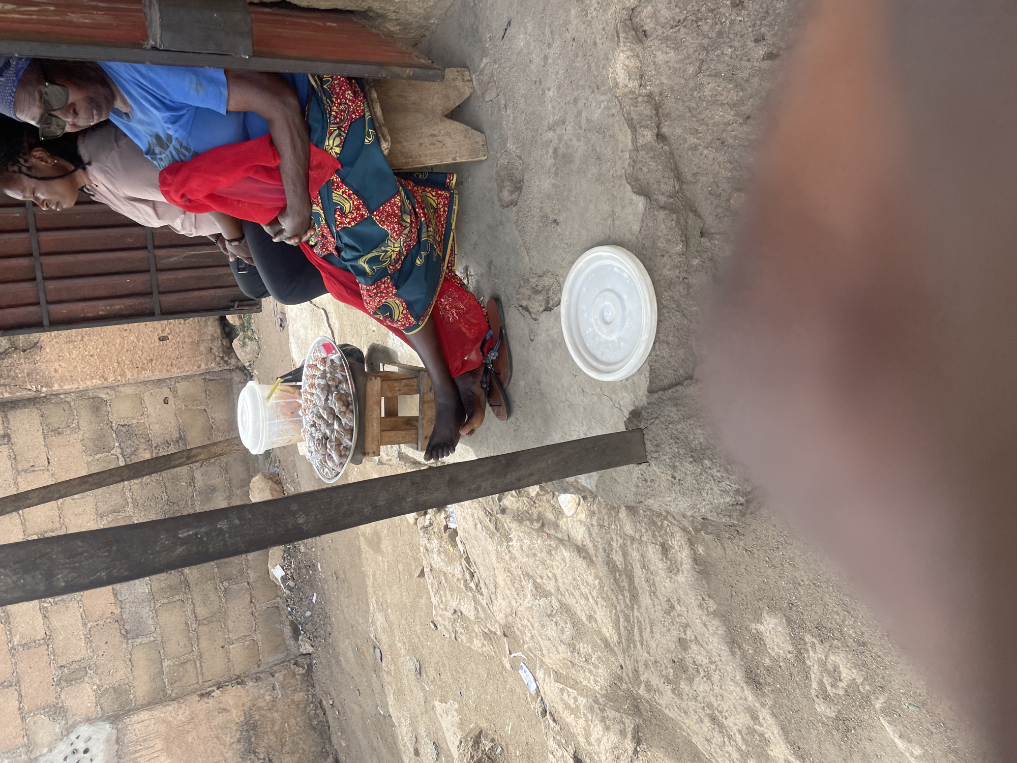 A partially obstructed, rotated aerial view shows a group of four people in an urban setting, likely in Jos, Nigeria. The people are seated in a makeshift arrangement using what appears to be a bench and small wooden stools. One person is wearing a blue shirt, another a light pink shirt, and a third is wearing a colorful patterned fabric. They appear to be engaged in a quiet activity or rest. Nearby, a shallow, circular container holds what may be food items or snacks. A white lid or container sits on the ground, suggesting the items might be for sale. The surrounding environment is a worn, concrete or paved area with walls made of cinder blocks. A metal beam crosses part of the image, causing further obstruction. The scene overall has a casual, street-side vibe.