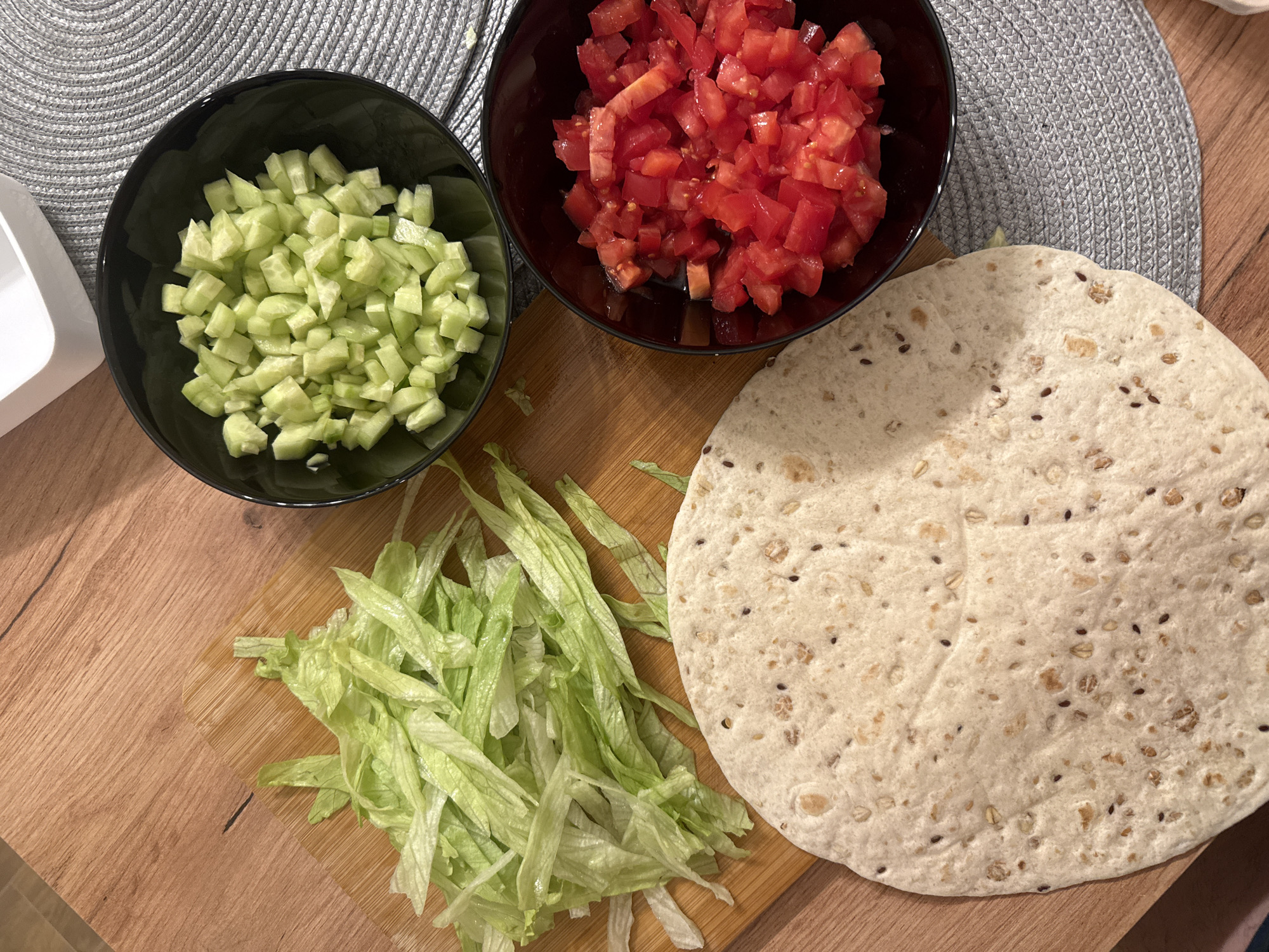 An overhead shot displays an assortment of fresh ingredients laid out on a light wooden surface, likely for meal preparation in a kitchen or dining setting.
In the upper left, a black bowl is filled with uniformly diced cucumbers. To its right, another black bowl contains brightly red, diced tomatoes. Below these bowls, on a small wooden cutting board, rests a pile of finely shredded green lettuce.
To the right of the lettuce and overlapping the cutting board, a large, round tortilla is visible. It has a light, possibly whole-grain appearance, with various seeds (such as flax seeds) and oat flakes visibly embedded in its surface. A grey, circular woven placemat is partially visible in the upper right and left corners, underneath the bowls and tortilla.
The scene suggests the activity of preparing a fresh meal, such as wraps, tacos, or a salad, with all the components ready for assembly. The lighting is even, indicating an indoor setting. No people or text are visible, and the specific time of day is not discernible. The overall impression is one of fresh, healthy ingredients arranged neatly for cooking or immediate consumption. The location context is Varna, Bulgaria, though this is not visually evident in the image itself.