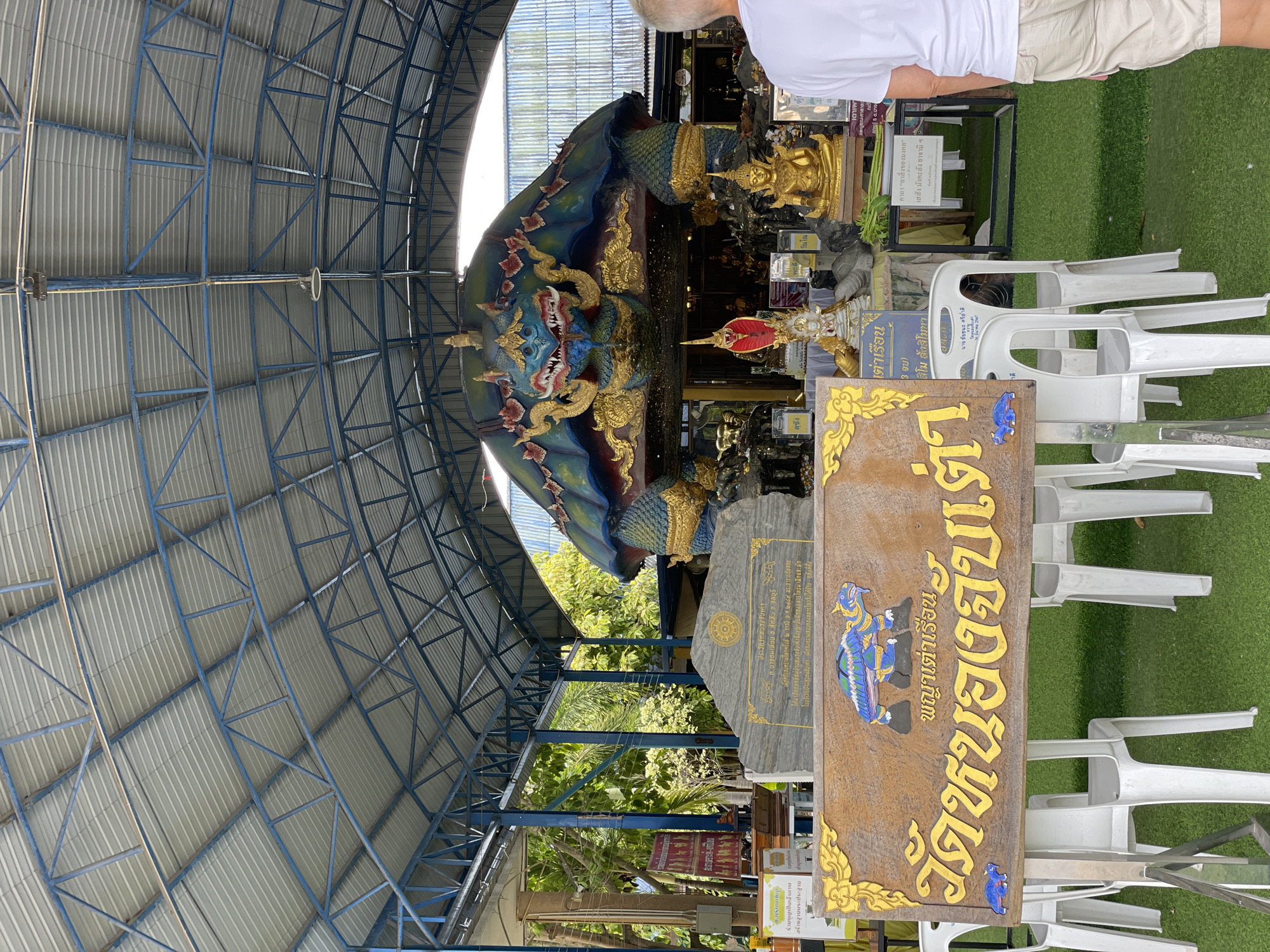 In Tambon Na Chom Thian, Thailand, an outdoor shrine features a large, colorful sculpture of a mythical turtle with a dragon's head, serving as the centerpiece. The structure is covered by a metal-framed, corrugated metal roof, supported by blue beams. In front of the sculpture, a wooden sign with Thai script and decorative carvings of turtles is displayed on a stand. The ground is covered with artificial turf. Several white plastic chairs are arranged in front of the sign and the shrine. In the foreground, a person wearing a white t-shirt and beige shorts stands facing away from the camera, looking towards the shrine. Behind the turtle sculpture, various statues and offerings are visible, including a golden Buddha statue and a red serpent figure. The environment suggests a public religious or tourist site.