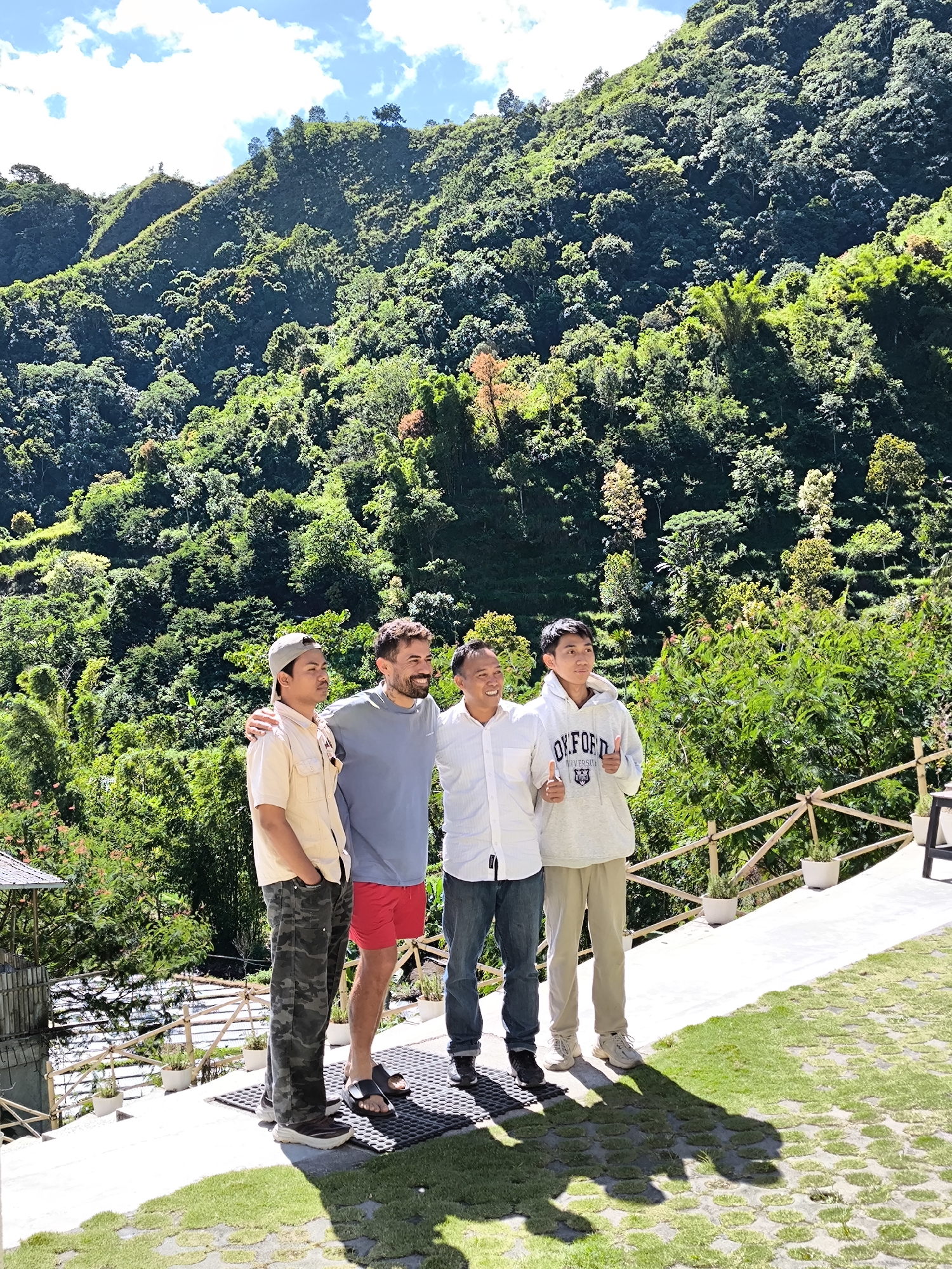 Four men stand together outdoors. The man on the left wears a tan button-down shirt, camouflage pants, and a cap. Next to him, a man with a beard wears a gray shirt and red shorts. The third man wears a white shirt and jeans. The man on the right wears a gray hoodie with the word "OXFORD" on it and tan pants. All men are smiling. The two men in the middle have their arms around each other. The man on the right is giving a thumbs up. They are standing on a stone walkway. In the background, there is a lush green mountain covered with trees. The sky is blue with some white clouds. They are in Sembalun Bumbung, Indonesia. A wooden fence and some potted plants are visible near the edge of the walkway. A small structure is seen behind the man on the left.