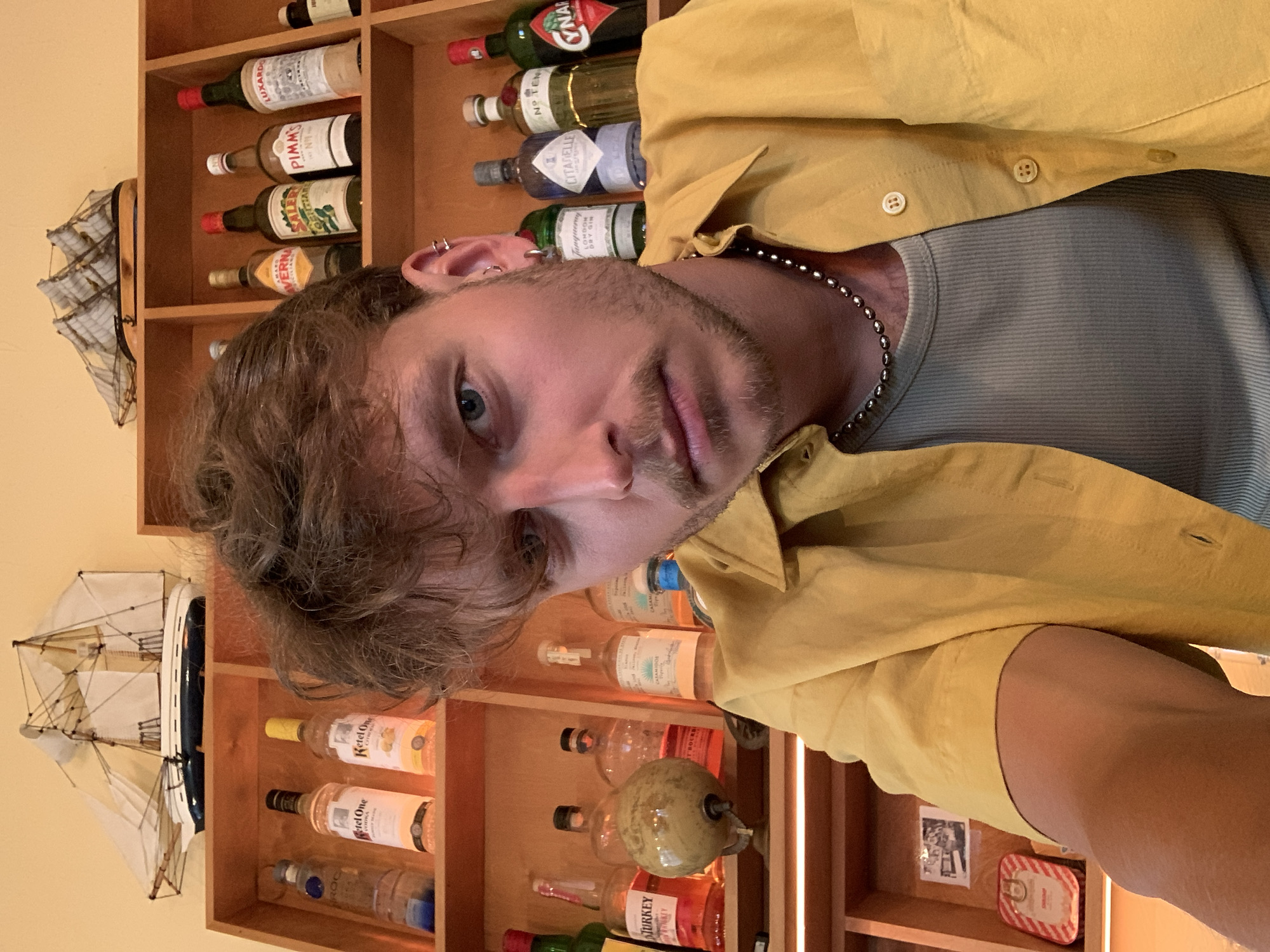 The image presents a head-and-shoulders portrait of a young man, likely a selfie, positioned against a background of wooden shelves filled with various bottles of alcohol. The man has light brown, slightly wavy hair, a short beard, and blue eyes, and is looking directly at the camera with a neutral to slightly pensive expression. He is dressed in a yellow collared shirt worn over a light grey ribbed t-shirt and accessorized with a dark beaded necklace and multiple ear piercings on his left ear.
The setting appears to be an indoor space, possibly a home bar or a dedicated liquor cabinet, with light-colored wooden shelves built into a yellow-painted wall. The shelves are extensively stocked with an assortment of spirits and liqueurs. Visible bottle labels include "LUXARDO," "PIMM'S No.1 CUP," "SALER'S" (likely Gentiane), "VERNA" (likely Amaro Verna), "Ketel One" vodka, "TANQUERAY LONDON DRY GIN," "CITADELLE," "NO TEN" (likely Tanqueray No. Ten Gin), "CYNAR," and "TURKEY" (likely Wild Turkey bourbon). A decorative model sailboat with white sails is displayed on a shelf to the left, and a small red and white striped tin is visible on a lower shelf. A warm, glowing light fixture illuminates the right side of the shelving unit. The location context indicates this scene is in Tel-Aviv, Israel.