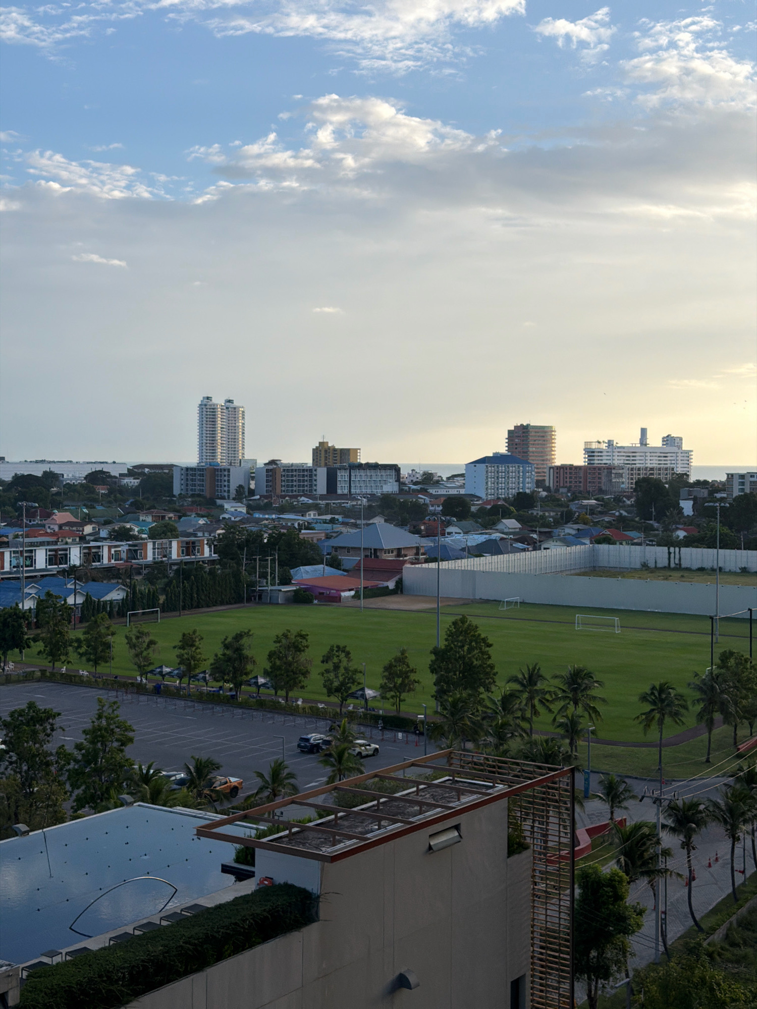 The image captures a wide aerial view of a coastal urban area, likely Hua Hin City Municipality, Thailand. In the foreground, a portion of a modern building is visible, featuring a swimming pool with dark water and a wooden lattice structure extending upwards. Adjacent to this, a parking lot is partially visible with several cars.
The mid-ground showcases a large, well-maintained green sports field, possibly a soccer pitch, enclosed by a tall, light-colored fence. Two white soccer goals are placed on the field. A paved running track encircles the field. A line of palm trees runs along the edge of the field, bordering a road with a few orange traffic cones. Further beyond the field, a dense residential area with houses of varying architectural styles and colors is spread out.
In the background, a cluster of taller buildings, including modern high-rises and mid-rise apartments, dominate the cityscape. The sea is visible in the distance, with a calm horizon. The sky is a mix of blue and scattered white clouds, with a soft, diffused light suggesting either early morning or late afternoon. The overall scene depicts a developed urban environment with recreational facilities, residential areas, and a coastal proximity.
