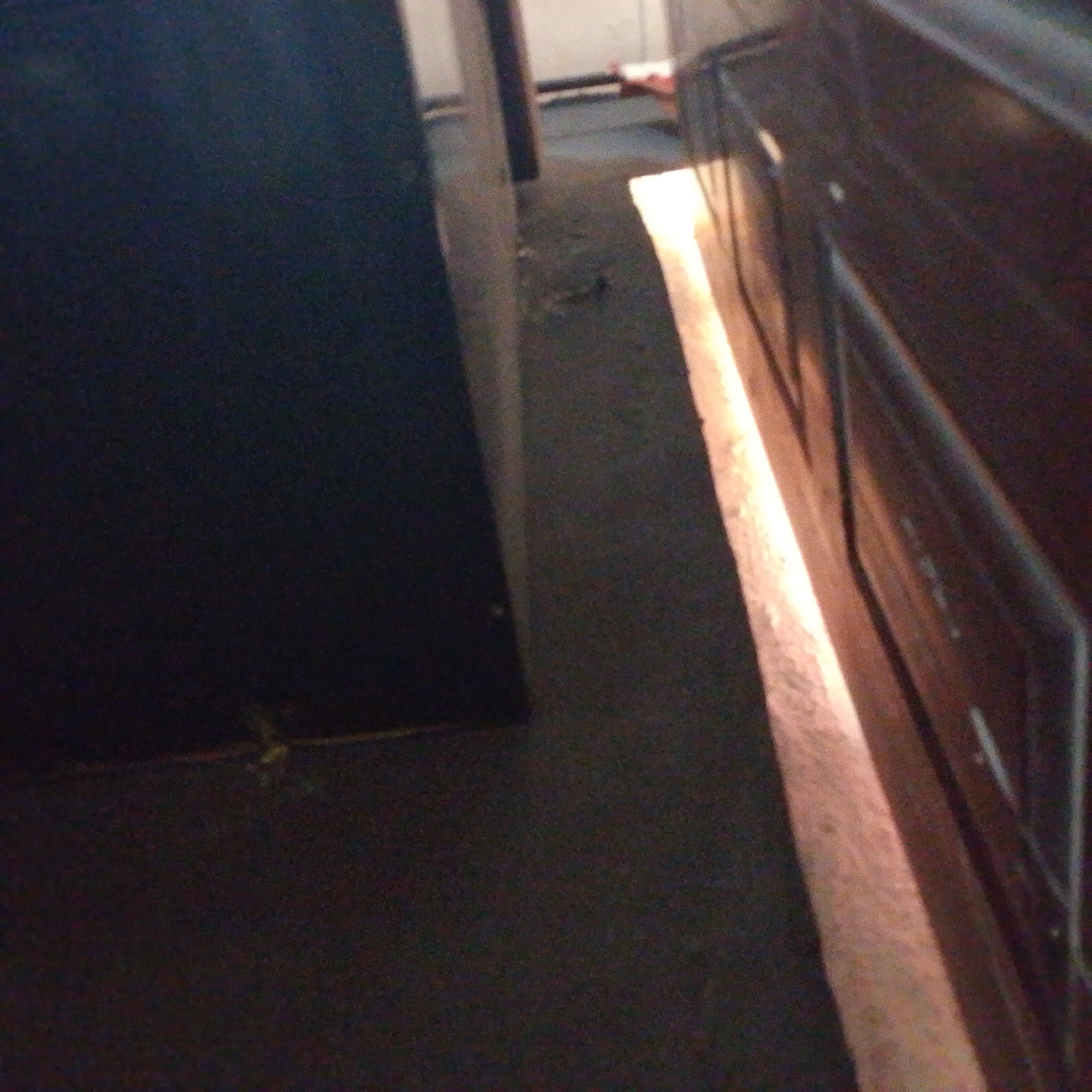 The image shows a narrow, dimly lit hallway or corridor with dark flooring and dark walls. A bright, light source illuminates a section of the floor and a portion of the right wall, creating a sharp contrast. The left side of the image is dominated by a large, dark, rectangular object, possibly a piece of furniture or a wall. The right side features a wooden surface with visible grain and some vertical lines, suggesting it might be a cabinet or a door. The floor appears to be carpeted or covered in a dark material, with some debris or shadows visible. The overall impression is one of a confined, utilitarian space, possibly a service area or a back corridor. The presence of the bright light suggests it might be a light fixture or light coming from an adjacent room. The image does not contain any discernible elements that would allow for specific identification of its location within Jalingo, Nigeria.