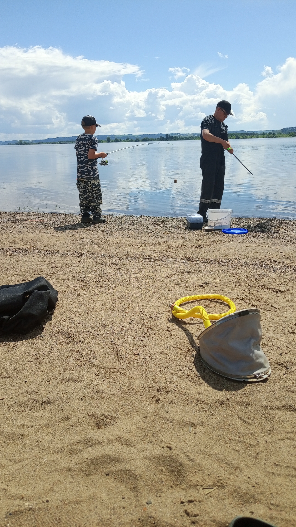 This image depicts an outdoor fishing scene at a wide body of water in Шолья, Russia, under a partially cloudy blue sky during daylight hours.
Two male individuals are positioned near the water's edge, engaged in fishing. On the left, a young boy, estimated to be between 7-10 years old, wearing camouflage attire and a dark baseball cap, holds a fishing rod with a spinning reel, directing it towards the water. To his right and slightly behind him, an adult male, wearing dark clothing, glasses, and a dark baseball cap, also holds a fishing rod, appearing to adjust its line or bait. A small, brown cylindrical bobber floats in the water between the two individuals.
On the sandy shore to the right of the adult male, a dark rectangular container with a grey lid, a white plastic bucket with a blue lid, and a black fishing net are visible near the water's edge. In the foreground on the sandy ground, a crumpled black fabric item lies on the left. To its right, a coiled yellow object, resembling a hose or rope, with a visible metal fitting, is positioned next to a light-colored bucket-like object with a flared rim, possibly a net or container.
The terrain consists of light brown sand mixed with pebbles near the water. The water is calm, reflecting the sky. The distant horizon features low-lying green land or hills beneath a sky populated by significant cumulus cloud formations.