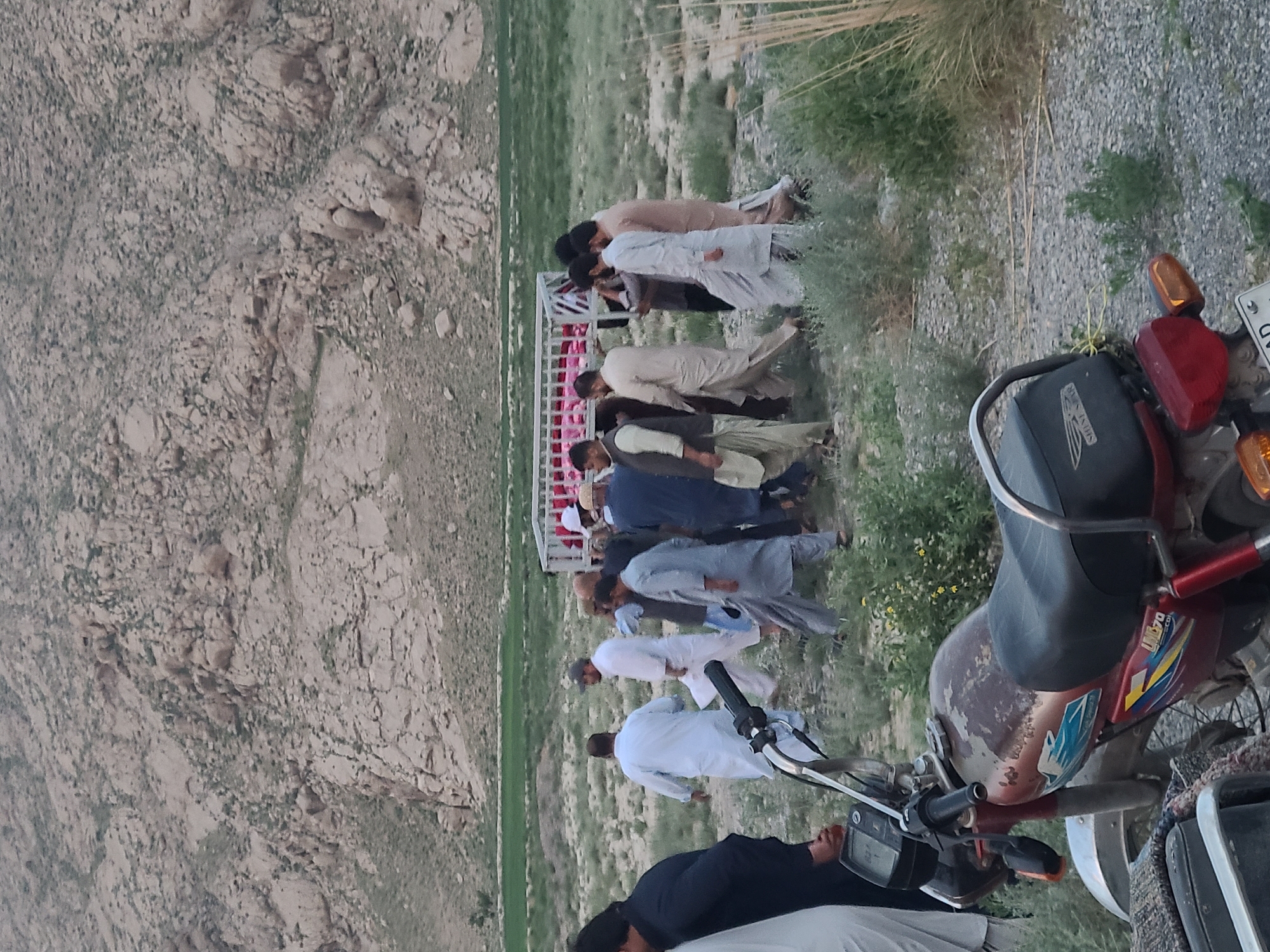 A group of approximately ten to fifteen men, dressed in traditional Pakistani attire such as shalwar kameez, are depicted in an outdoor, arid landscape, likely in Quetta City Tehsil, Pakistan. Most men wear light-colored clothing (white, cream, light brown, light blue), with one individual in the foreground wearing dark attire.
The men appear to be walking in a loose procession or gathering, moving from left to right across the frame. Behind some of the men, a white metal structure with pink accents, possibly part of a cart or a temporary shelter, is visible.
The setting is characterized by rugged, light-colored rocky mountains and hills in the background, indicative of a mountainous and dry region. The ground is covered with sparse, dry grass and gravel, with patches of greener vegetation further in the distance.
In the foreground on the right, a red and black motorcycle is parked. Visible text on the motorcycle includes "SHINE STAR" on the engine cover and "LOADER 70" on the fuel tank. A partial license plate shows "QC", which likely stands for Quetta.
The lighting suggests either late afternoon or early evening, with soft, diffused light and no harsh shadows. The overall atmosphere is quiet, and the men's expressions are not clearly discernible due to distance.