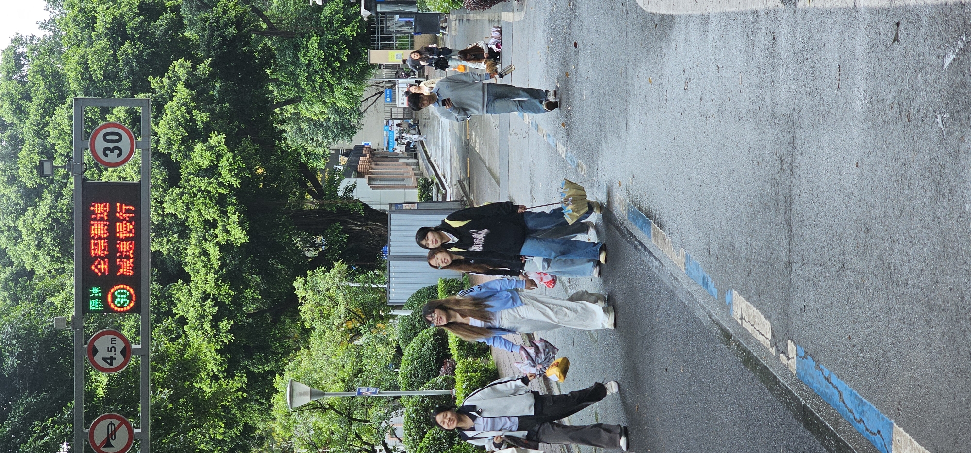 The image shows a street scene in the Yuzhong District of China. A group of people, mostly young women, are walking along the street. One is holding an umbrella. In the background, there are trees, a street sign with a speed limit of 30, and buildings. The street appears to be wet.