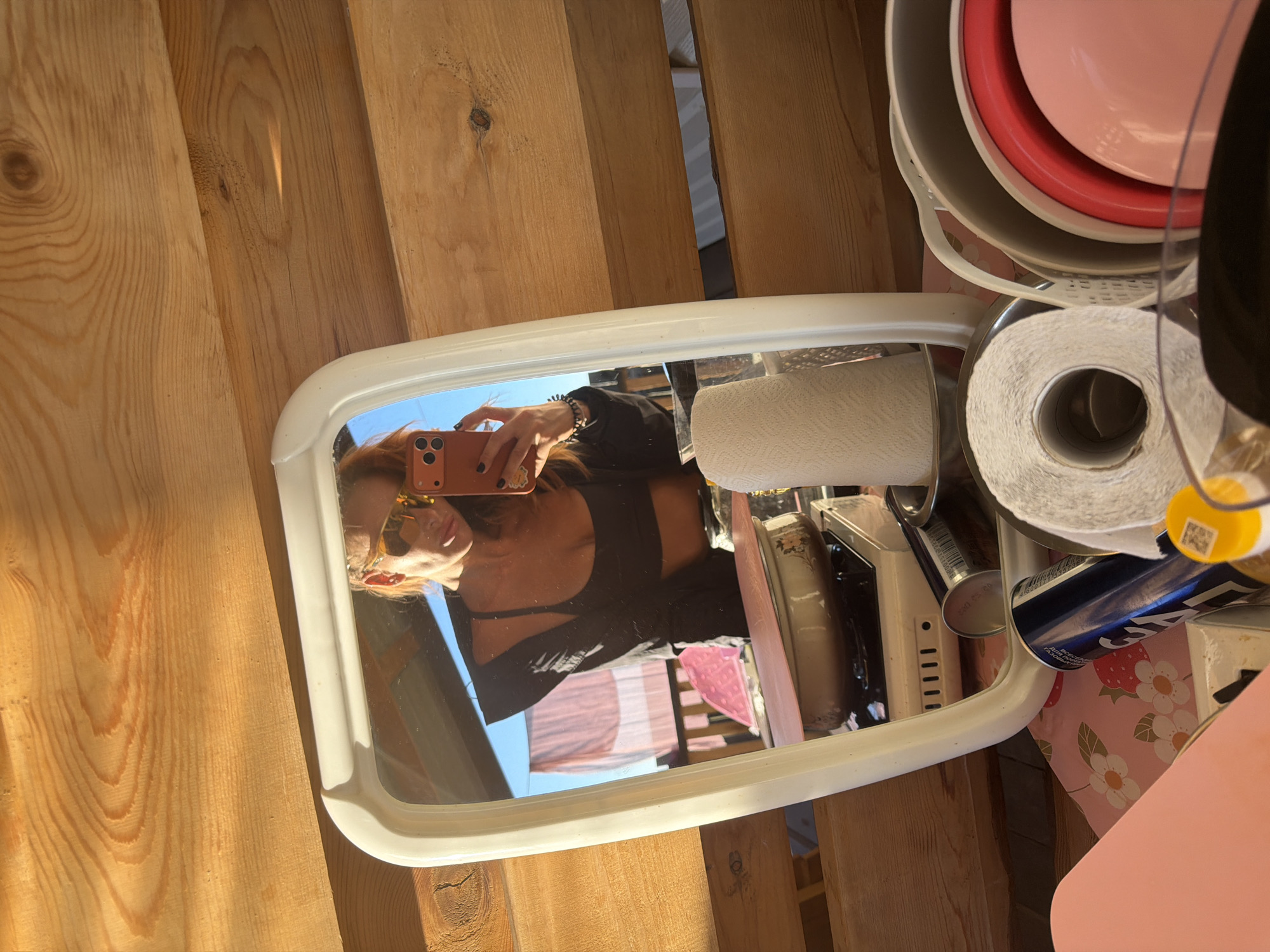 The image depicts a woman taking a selfie in a mirror. She is wearing sunglasses and a black top, and is holding an orange smartphone. In the mirror's reflection, a portable stove with a pot on it is visible, along with a roll of paper towels and other kitchen items. The background consists of wooden planks and a blue sky with some laundry hanging. The scene appears to be outdoors, possibly on a balcony or in a rustic setting. There is no identifiable information about the location in the city of Unknown, Russia.