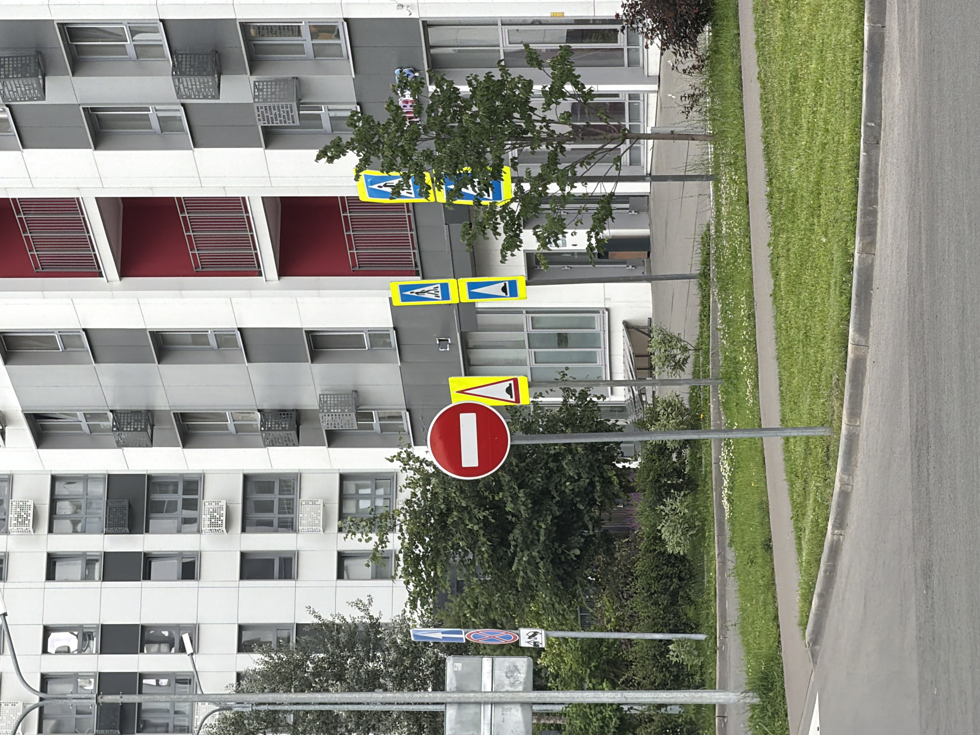 This media file shows a residential building with a row of trees and a sidewalk along a road. The building is modern, with a white facade and dark-colored balconies. The trees are lush and green, suggesting it is a warm season, likely spring or summer. The weather appears to be overcast, as there are no strong shadows. The scene is oriented vertically, suggesting the photo was taken with a phone in portrait mode.
Several traffic signs are visible. The most prominent is a red circular sign with a white horizontal bar, indicating a "no entry" or "do not enter" restriction. Above it, a yellow triangular sign shows a black silhouette of a vehicle with a directional arrow, likely indicating a turn restriction or a specific lane usage. Further down, there are signs indicating pedestrian crossings. One sign has two blue arrows on a yellow background, pointing in opposite directions, possibly indicating a two-way street or a crossing. Another sign below it has a blue arrow pointing left and a red circular sign with a diagonal bar across it, suggesting a prohibition.
The presence of multiple traffic signs, particularly those related to pedestrian crossings, suggests this is a public road in a residential area. The building appears to be in a well-maintained urban or suburban environment. There is no visible text other than on the traffic signs. The image captures a quiet moment, with no people or vehicles actively present.