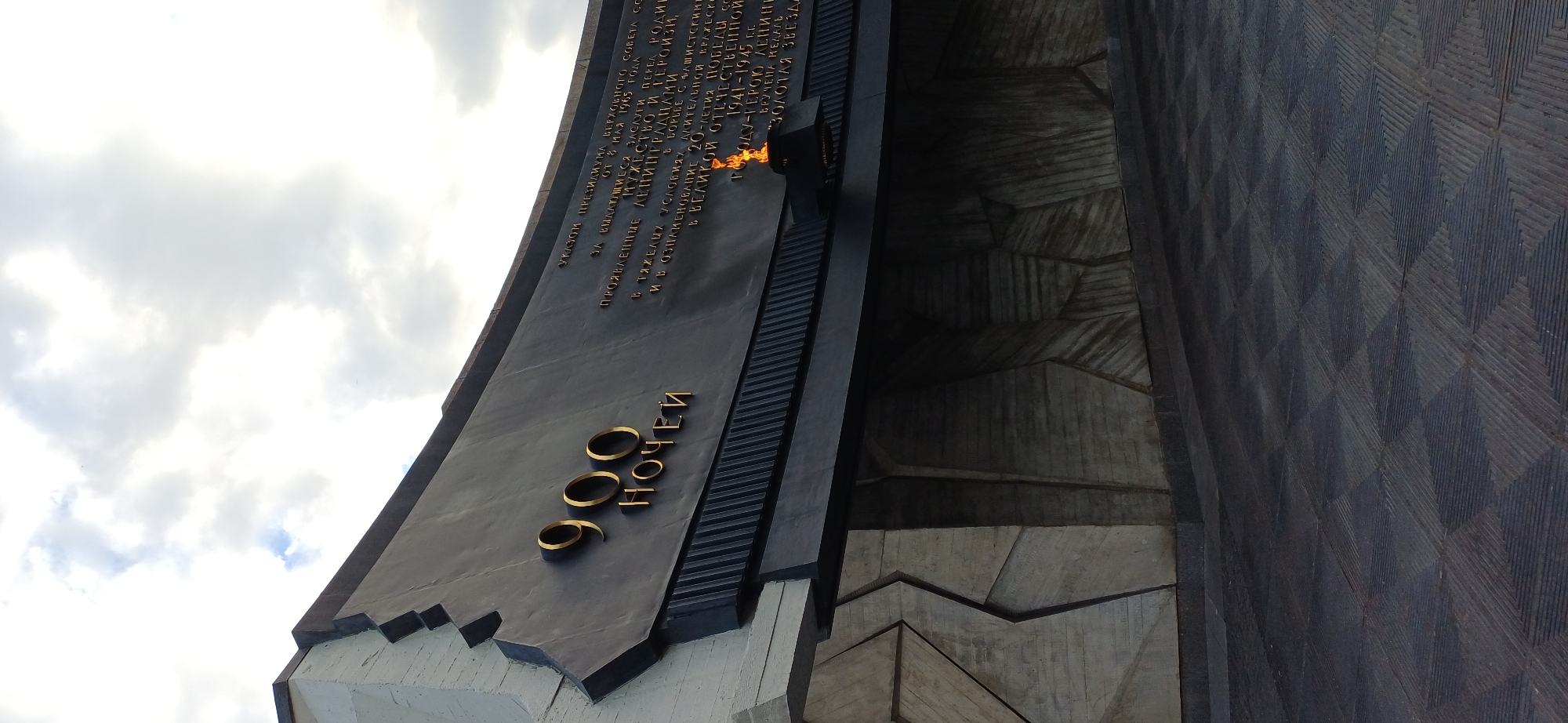 The image shows a monument with golden lettering and an eternal flame. The lettering on the dark grey curved surface reads "900 NOCHEЙ" (900 Nights). To the right of this, there is a long inscription in Russian, which appears to be a decree or award. The monument is made of dark grey metal and concrete with a textured, angular design. The eternal flame burns in a metal receptacle in front of the inscribed text. The ground in front of the monument is paved with dark tiles arranged in a diamond pattern. The sky above is cloudy, with patches of blue visible. This monument is located at the Piskaryovskoye Memorial Cemetery in Saint Petersburg, Russia, dedicated to the victims of the Siege of Leningrad.
