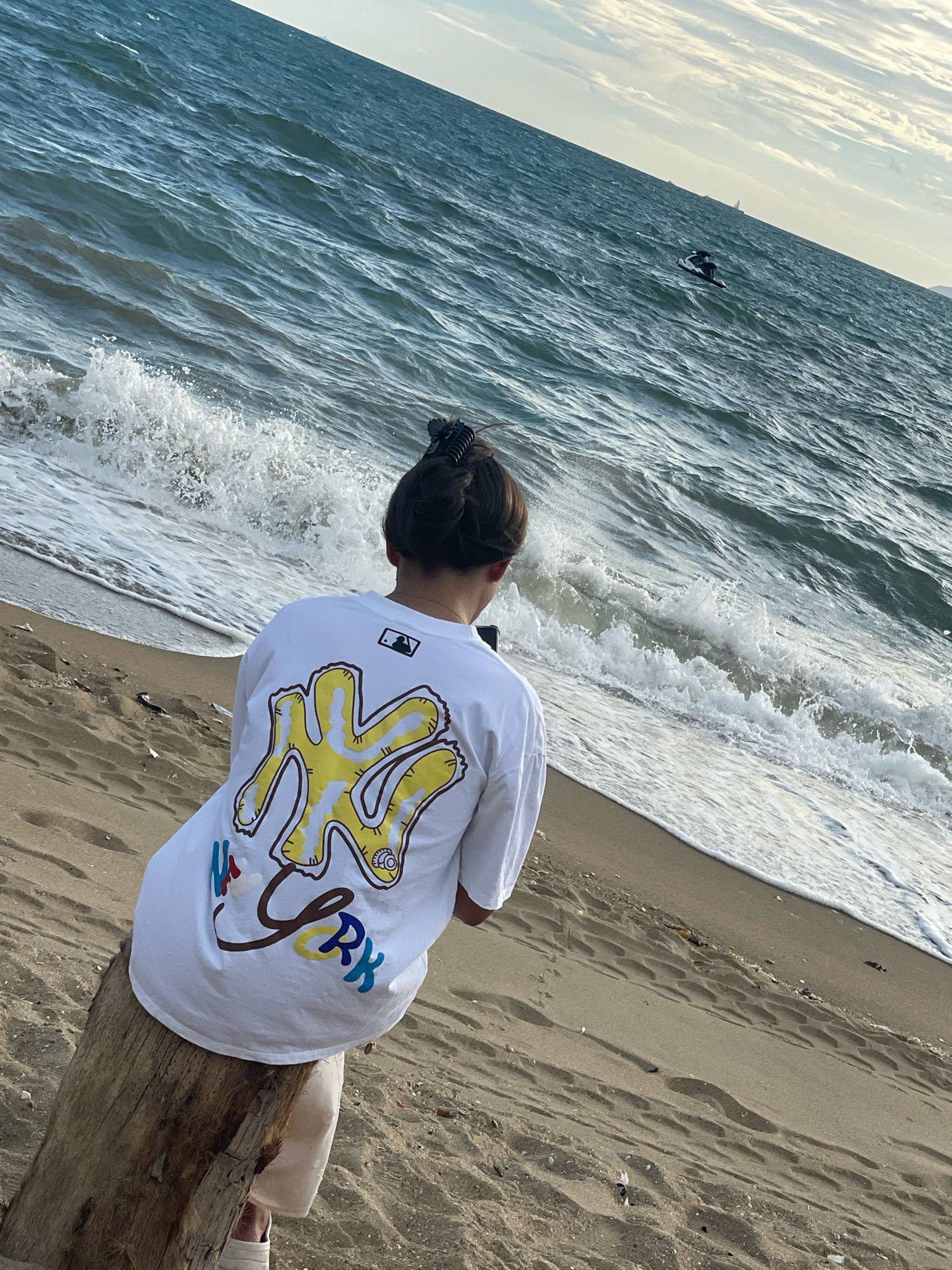 The image depicts a person sitting on a log on a sandy beach in Pattaya City, Thailand. The person is wearing a white t-shirt with a graphic design on the back and light-colored pants. They are facing away from the camera, looking out at the ocean. The sea is blue with waves breaking on the shore. In the distance, a jet ski is visible on the water. The sky above is cloudy. The sand is light brown and shows footprints.