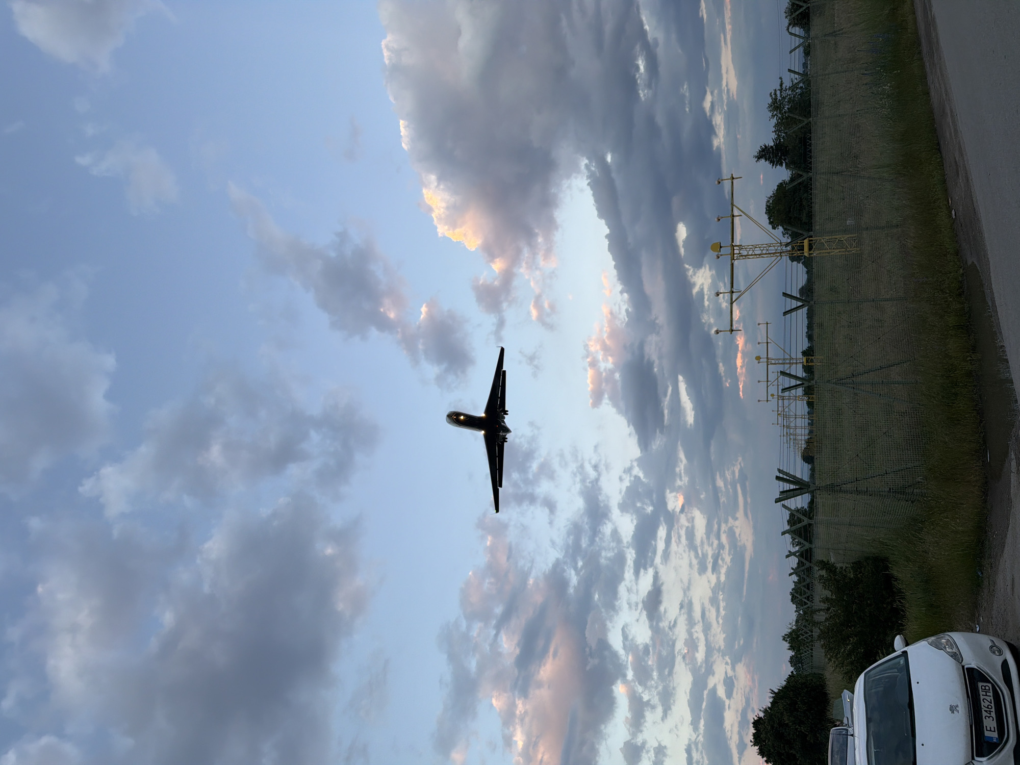 A low-angle, landscape-oriented image (originally portrait and rotated) captures an airplane flying low overhead against a dramatic, partially cloudy sky during dusk or dawn. The scene is set near an airport runway in Sofia, Bulgaria.
The primary subject is a dark silhouette of an airplane, likely a jet, positioned centrally in the upper half of the frame, flying from right to left. Its landing lights or internal cabin lights are visible as small, bright points on its underside. The sky displays a gradient of colors, from a pale blue closer to the horizon to darker, dense clouds, some of which are illuminated with warm orange and pink hues by the setting or rising sun.
In the midground, running along the right side of the frame, is a yellow-colored airport approach lighting system, consisting of support structures and lights. Below this, a tall wire fence stretches horizontally, separating a wild, grassy field from a paved surface, likely a road or service lane, in the foreground. Trees and dense foliage are visible behind the fence.
In the bottom right corner, the front section of a white Peugeot car is partially visible. The car's license plate, "E 3462 HB," is clearly legible, confirming its Bulgarian registration.
The image conveys the activity of an airplane either approaching for landing or taking off from an airport. The soft, ambient light and the colorful, cloud-filled sky create a serene yet dynamic atmosphere.
