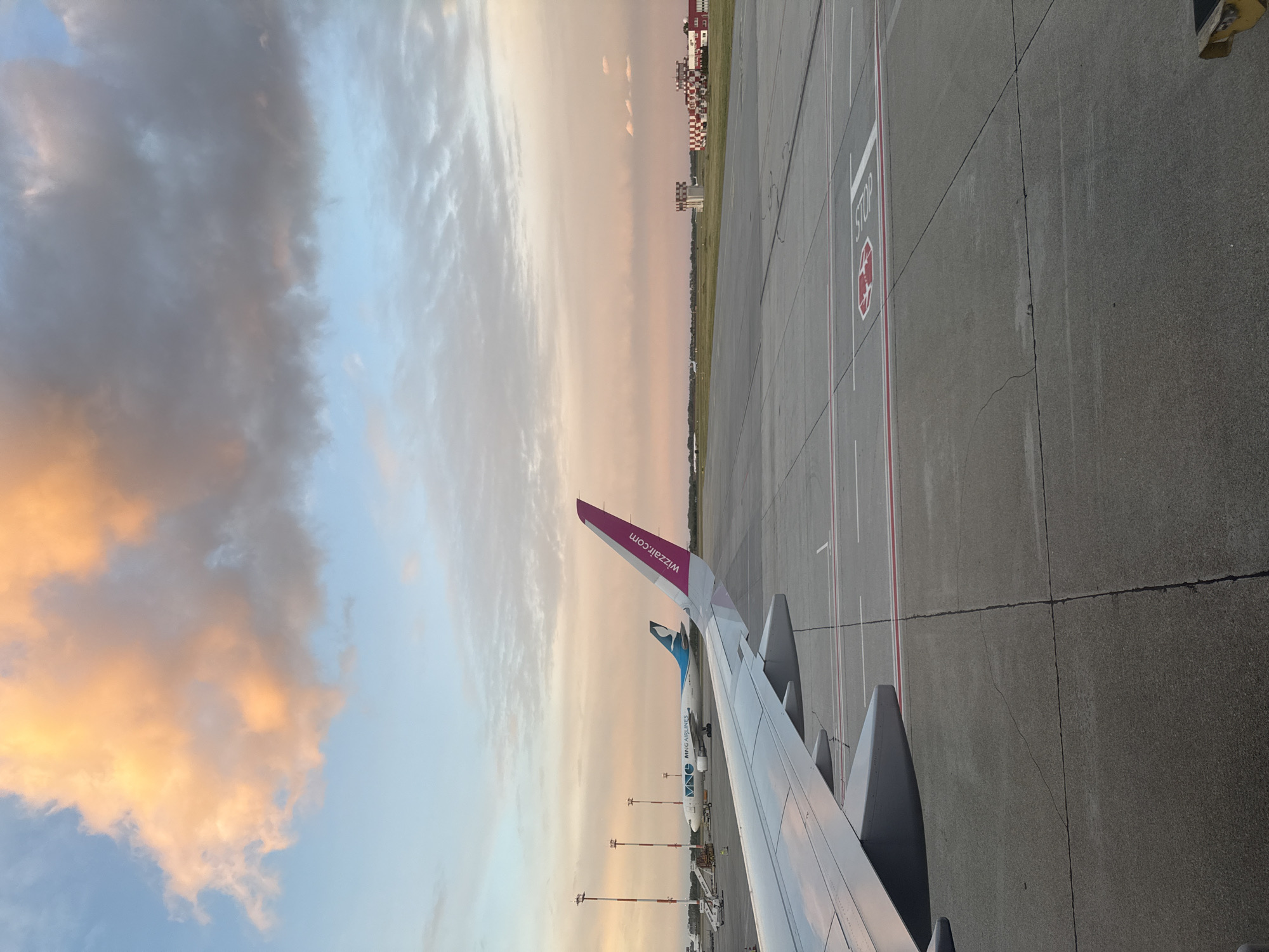 The image shows an airplane wing with the wizzair.com logo on the winglet, situated on an airport tarmac, possibly at Bratislava Airport in Slovakia. The sky is visible with a mix of blues, whites, and oranges, suggesting either sunrise or sunset. The tarmac is marked with lines and directions, including what appears to be a "STOP" sign. In the distance, there are buildings and another aircraft. No people are directly visible in the image.