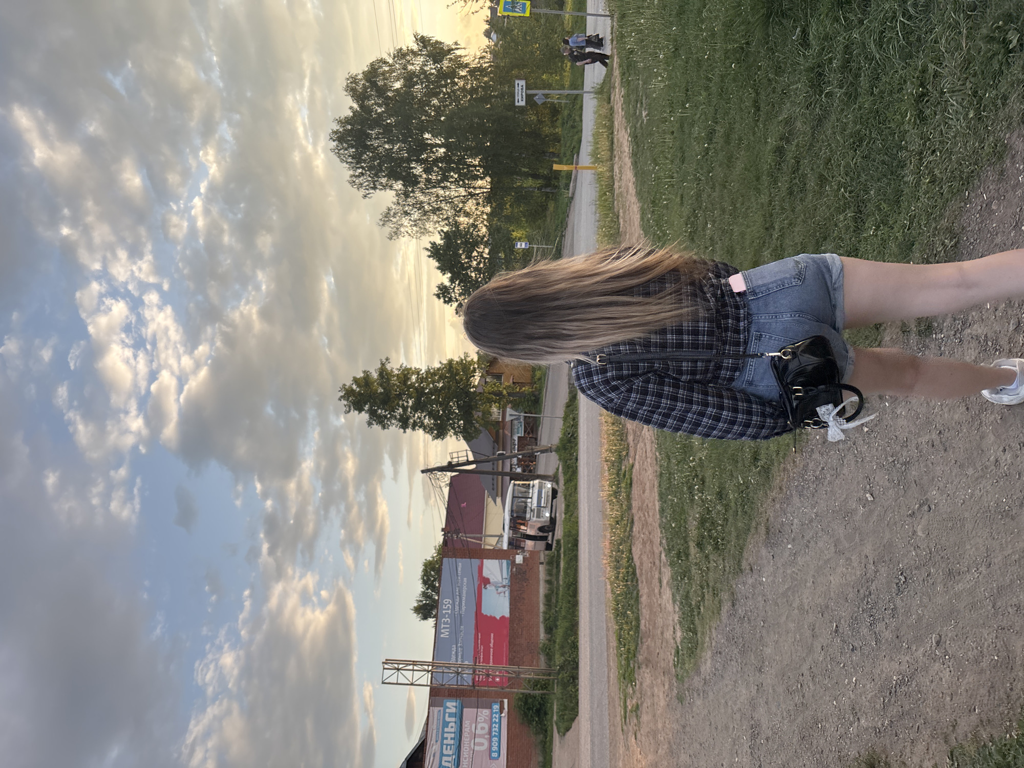 The image shows a young woman with long blonde hair, wearing a plaid shirt and jean shorts, walking along a dirt path. She is seen from the back. The path is bordered by grass on one side and buildings on the other. The buildings include a brick structure with a large advertising billboard. The billboard contains Russian text, including phone numbers and the text "MT3-159". Across the street is a white bus. In the background, there are trees and a cloudy sky with sunlight filtering through. The location is identified from the billboards as Фоки, Russia. In the distance, there are pedestrian crossing signs.