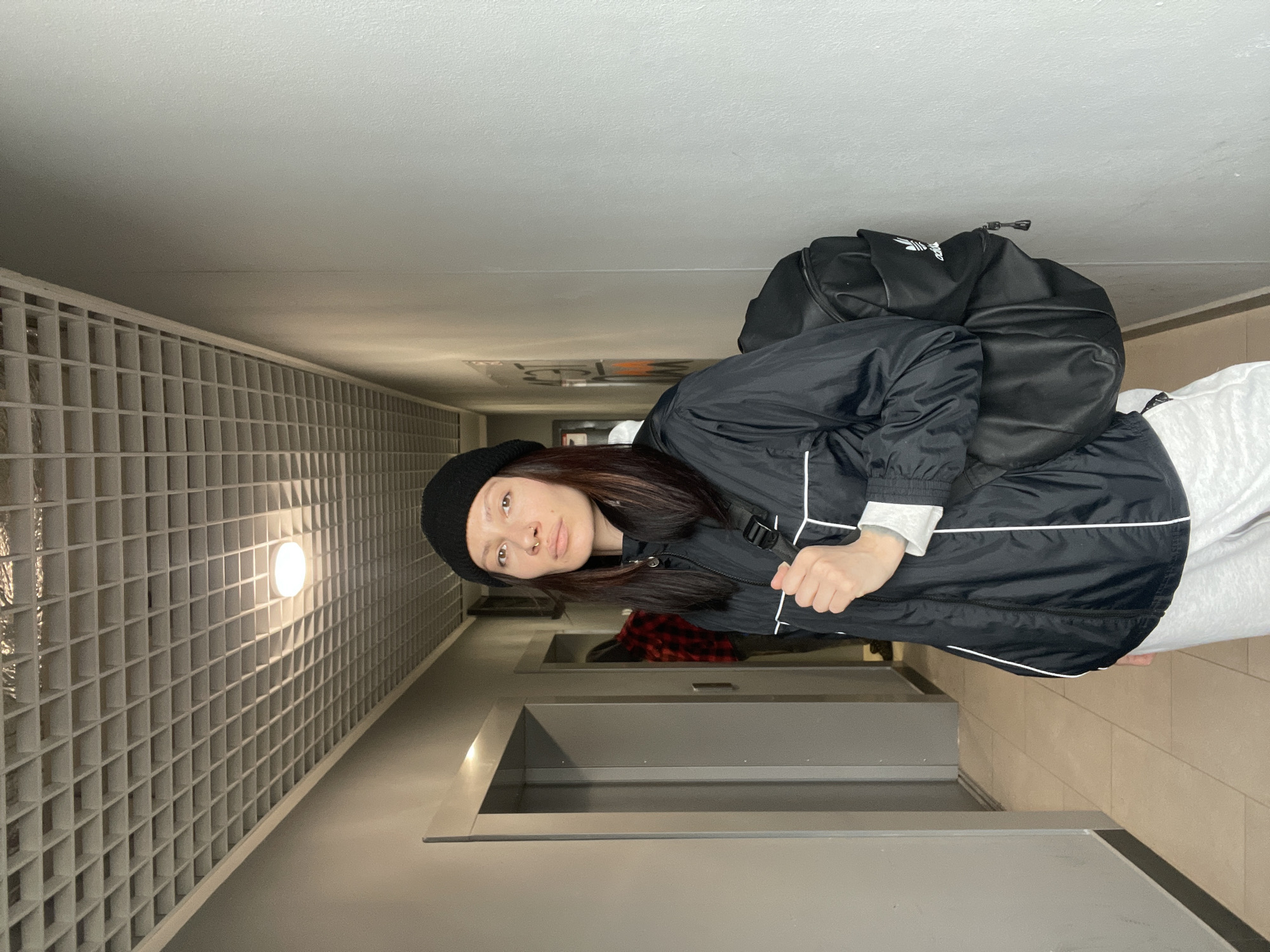 A woman wearing a black beanie, black jacket with white trim, and a black backpack stands in a narrow hallway. The hallway has a wall with a grid-like pattern on one side, a light fixture illuminating the grid. A door is visible at the end of the hallway. The image is taken from a low angle and rotated. The location is potentially in a building in Izhevsk, Russia.