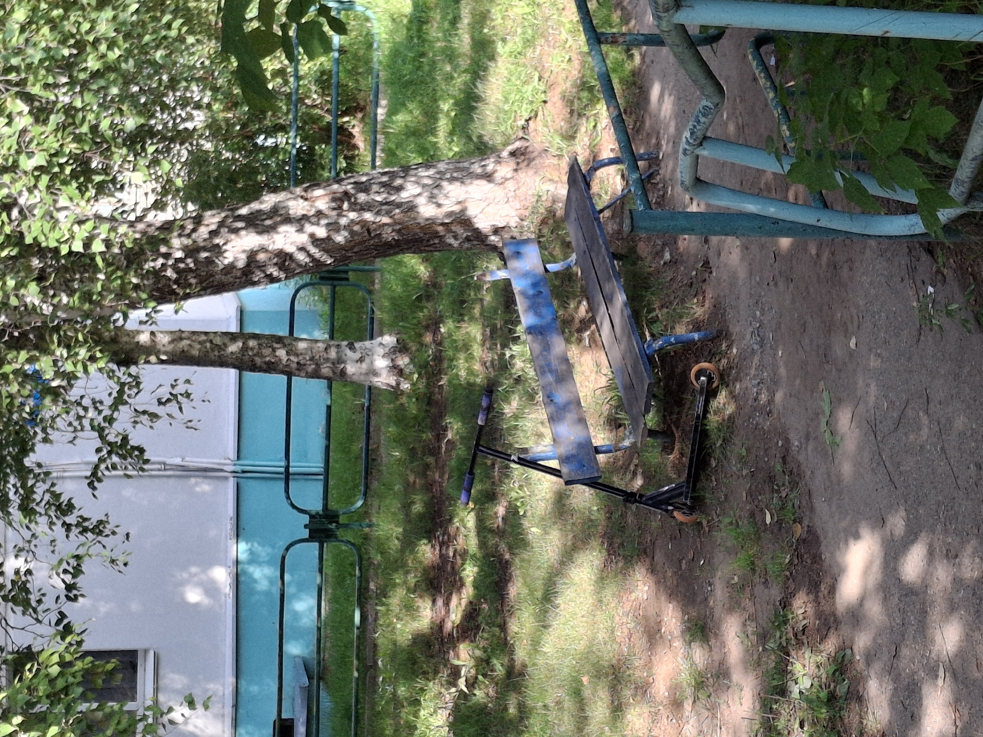 The image depicts a scene outdoors, likely in a residential courtyard in the city of Chaikovsky, Russia, based on contextual information. In the foreground, a black scooter with orange wheels is parked on a dirt path next to a weathered blue wooden bench. The bench is positioned under the dappled shade of trees, with its wooden planks showing signs of wear and tear. Behind the bench, a large tree trunk with textured bark is prominent. Further back, a portion of a light-colored building with a turquoise band runs along its base, suggesting it is a residential structure. A turquoise metal fence runs horizontally across the mid-ground, separating the courtyard area from a grassy expanse beyond. The environment is characterized by greenery, grass, dirt, and the presence of trees and a building, indicative of a common urban living space. The overall impression is one of a quiet, possibly abandoned, outdoor area.