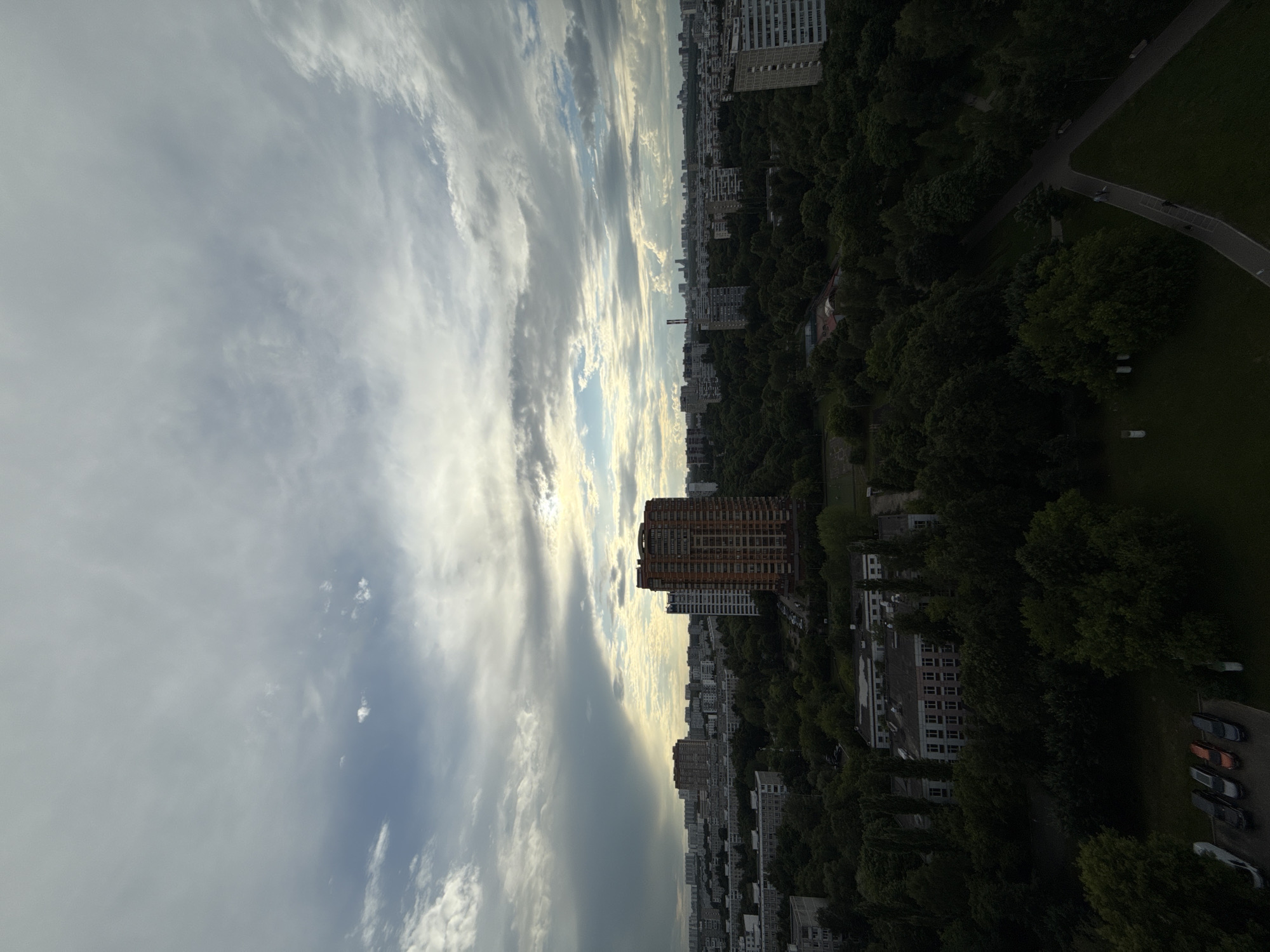 The image captures a wide aerial view of a residential district in Moscow, Russia, under a cloudy sky. The foreground is dominated by dense green trees, with a few buildings and a parking lot with several cars visible. A paved walkway with a couple of people can be seen in the lower right portion of the image. In the mid-ground, more apartment buildings of varying heights and architectural styles are interspersed with greenery. A prominent, tall, orange-brick apartment building stands out in the center-right. In the background, a sprawling cityscape of numerous buildings stretches towards the horizon, with a few taller structures visible in the distance. The sky is filled with a mix of dark and light clouds, with a sliver of sunlight breaking through in the upper right, suggesting late afternoon or early evening.