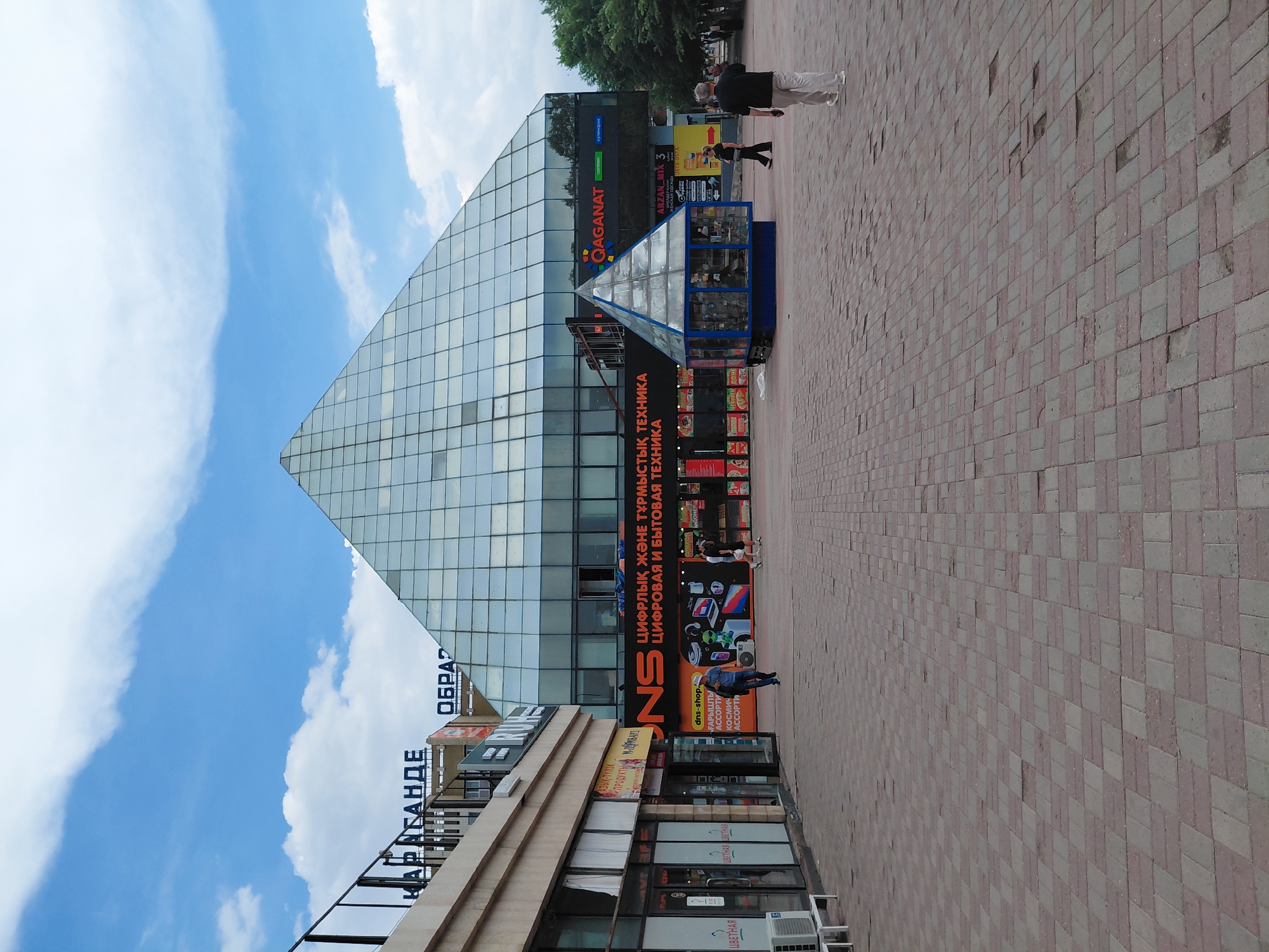 The image depicts an outdoor plaza with a large, modern, glass-pyramid-shaped building dominating the background. The plaza is paved with reddish-brown and gray interlocking bricks. To the left, a beige building with multiple storefronts and dark-tinted windows is visible. Above one of these storefronts, blue Cyrillic lettering reads "ҚАРАҒАНДЫ" which translates to "QARAGANDY". To the right of the pyramid building, a smaller, glass-enclosed kiosk with a blue base is situated.
Several people are present in the scene. In the foreground, a man in a dark shirt and light-colored pants walks away from the viewer towards the right. Further into the plaza, a man in a blue shirt and jeans walks towards the left, appearing to be on his phone. A woman in black pants and a dark top walks to the left, and a man in a dark shirt and lighter pants walks towards the pyramid building. Two individuals are walking together in the distance on the right side of the plaza, near a line of trees.
The pyramid building has a glass facade with a grid pattern. Below the pyramid, an orange sign with black Cyrillic lettering is visible, reading "DNS SHOP". Smaller signs above it indicate services. The sky is blue with scattered white clouds. Trees are visible behind the pyramid building on the right. The overall scene suggests a public area, likely a commercial or administrative center in Qarağandı, Kazakhstan.