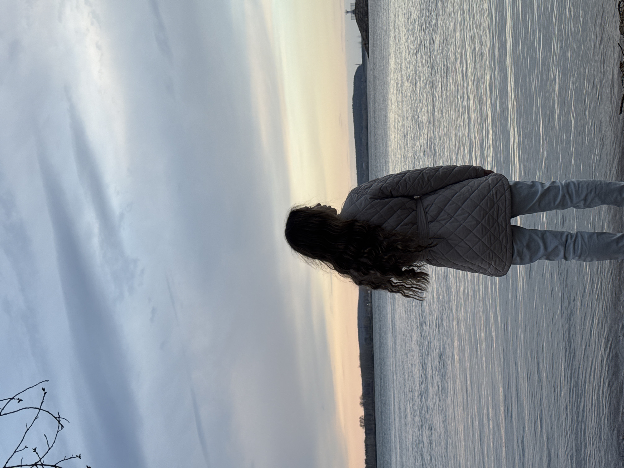 A young person, seen from the back in a side profile, stands at the edge of a large body of water, gazing towards the horizon. She has long, dark, wavy hair and is wearing a light-colored quilted jacket and light pants. The sky dominates the upper portion of the frame, displaying a gradient from muted blues and greys overhead to a pale yellow-orange hue near the horizon, indicative of either sunrise or sunset. The expansive body of water, likely a river or reservoir, features subtle ripples on its surface, reflecting the soft light. In the far distance, a faint shoreline with low hills and trees can be discerned, and a tall, slender structure, possibly a lighthouse or tower, is visible on a distant landmass. In the lower left foreground, bare tree branches with small buds suggest an early spring or late winter season. The atmosphere appears calm and contemplative. This tranquil scene is set in Chaikovsky, Russia. There is no visible text.