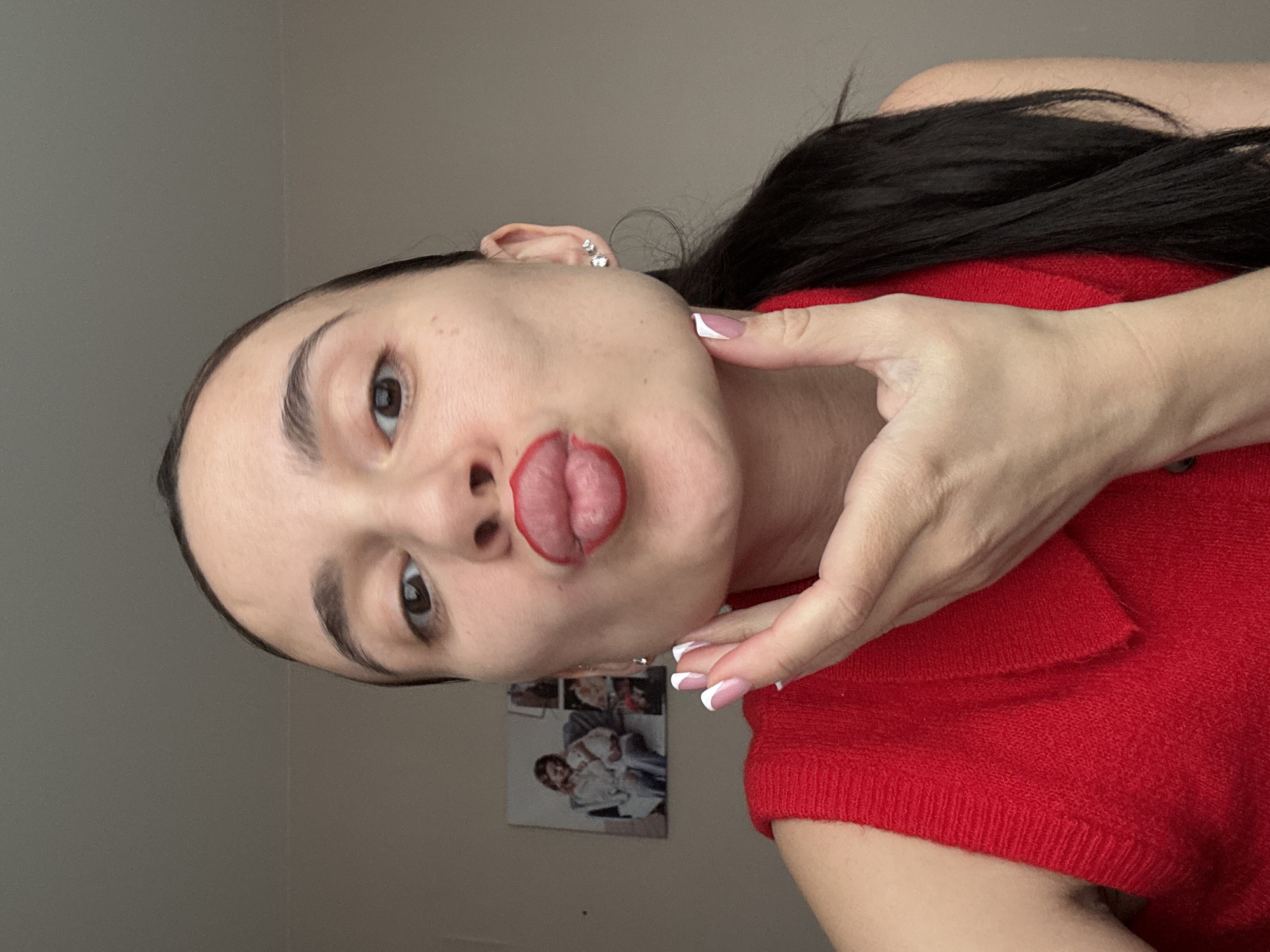 A young woman with dark hair and fair skin is pictured indoors. She's wearing a red top. Her lips are puckered and have red lipstick applied around the outline. She has manicured nails with white tips. Her right hand is positioned near her face. Behind her is a wall with a picture on it. The location is likely in a residential setting, not specifically identifiable as the city of Chaikovsky, Russia without additional context or identifying features in the image.