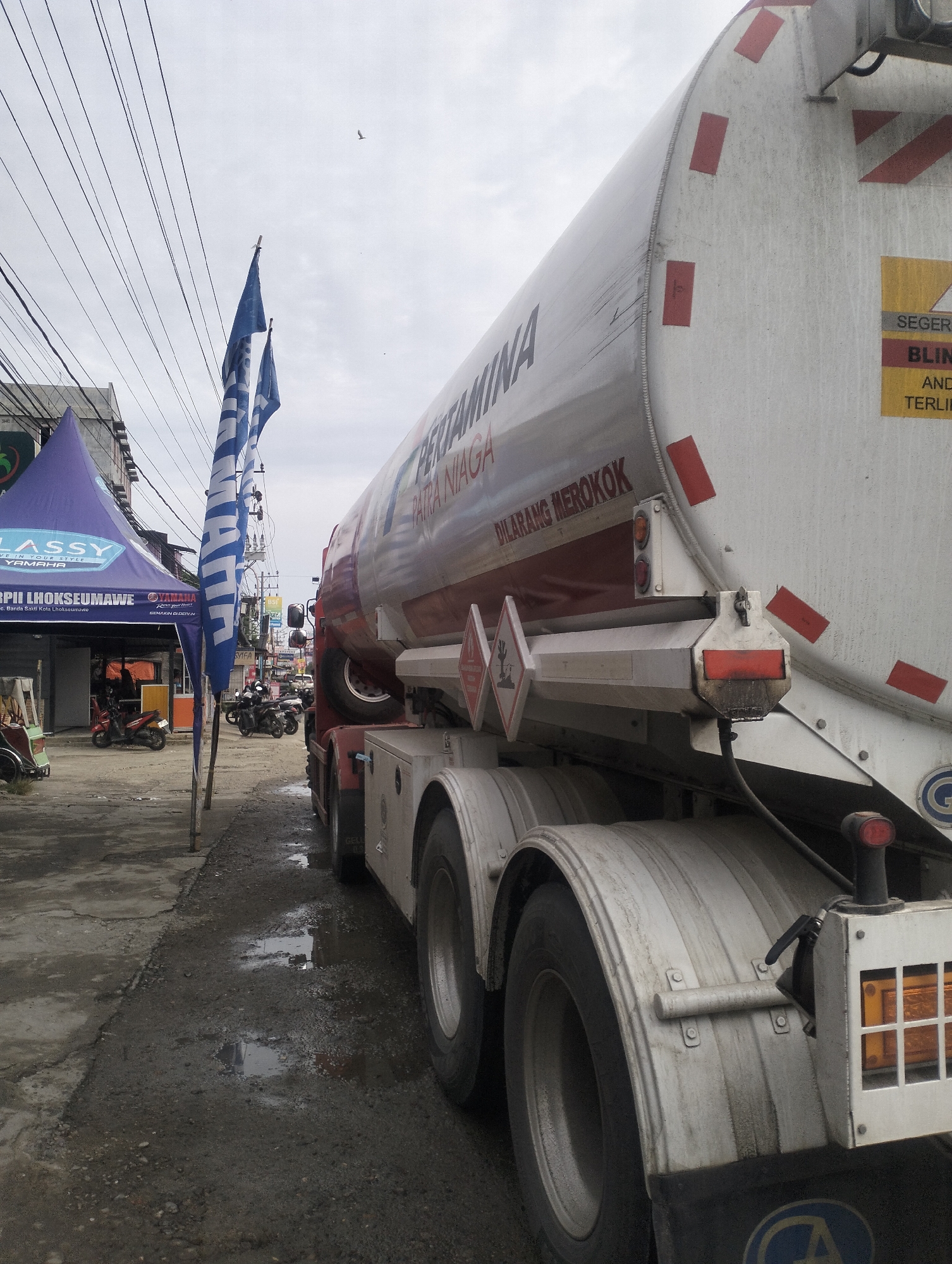 The image depicts a street scene in Lhokseumawe, Indonesia, with a large white and red fuel tanker truck dominating the foreground. The truck, identified with the logo "PERTAMINA," is parked on a wet, dirty asphalt road, suggesting recent rain or water spillage. Two blue and white striped flags, likely promotional or event-related, are planted vertically on the left side of the road, in front of a purple tent. The tent has "CLASSY" and "YAMAHA" logos visible, indicating a business or event location. The sky is overcast. In the background, a few motorcycles are parked and some people can be seen near the tent. Power lines run overhead. The overall impression is of a typical urban street environment in Indonesia, likely a commercial area.