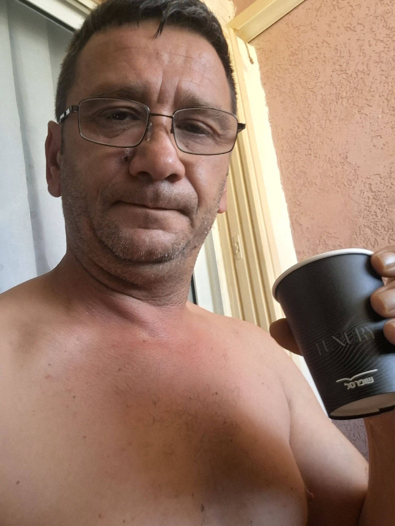 A man with glasses and stubble is taking a selfie outdoors, holding a black paper cup with a white rim. He is shirtless and his skin appears red and possibly sunburned on his chest and neck. The background shows a textured, peachy-colored wall and a light-colored vertical element which may be a door or window frame. The writing on the cup is partially visible and appears to be in Hebrew. The scene suggests an outdoor setting, possibly a residential area.
