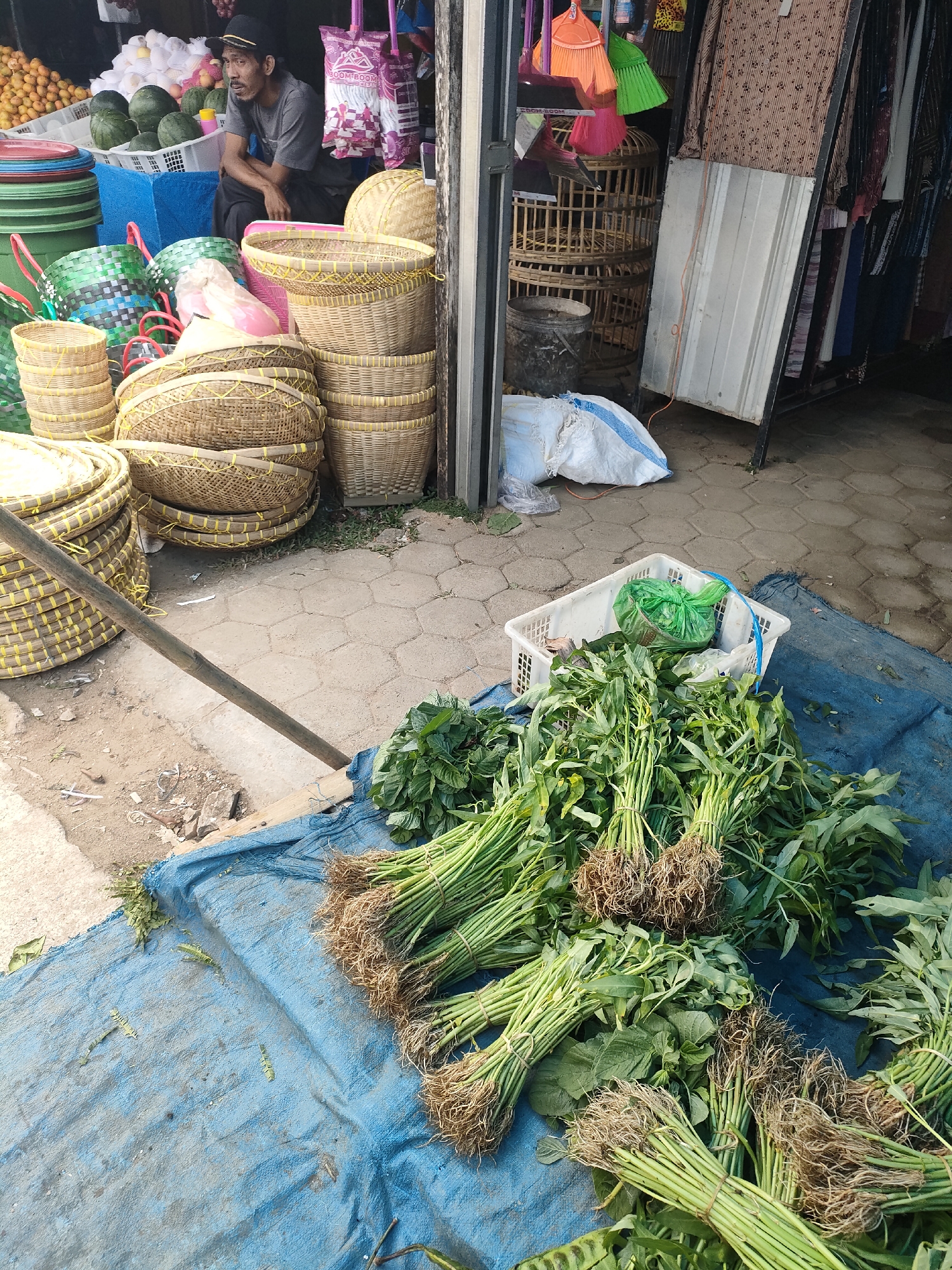 The image depicts an outdoor market stall in Sukoyoso, Indonesia. A man wearing a dark baseball cap and a gray t-shirt sits on a black plastic stool amidst a variety of goods. To his left, there are stacked woven baskets of various sizes, some with yellow detailing. Behind him, there are also plastic containers filled with fruit, watermelons, and stacked green and pink plastic baskets. Further to the right, hanging items like brooms and clothing are visible, along with bird cages. In the foreground, several bundles of green vegetables with visible roots are laid out on a blue tarp. A white plastic crate containing a green plastic bag is also present on the tarp, near the vegetables. The ground is paved with hexagonal tiles and dirt. The scene appears to be a typical street market environment.