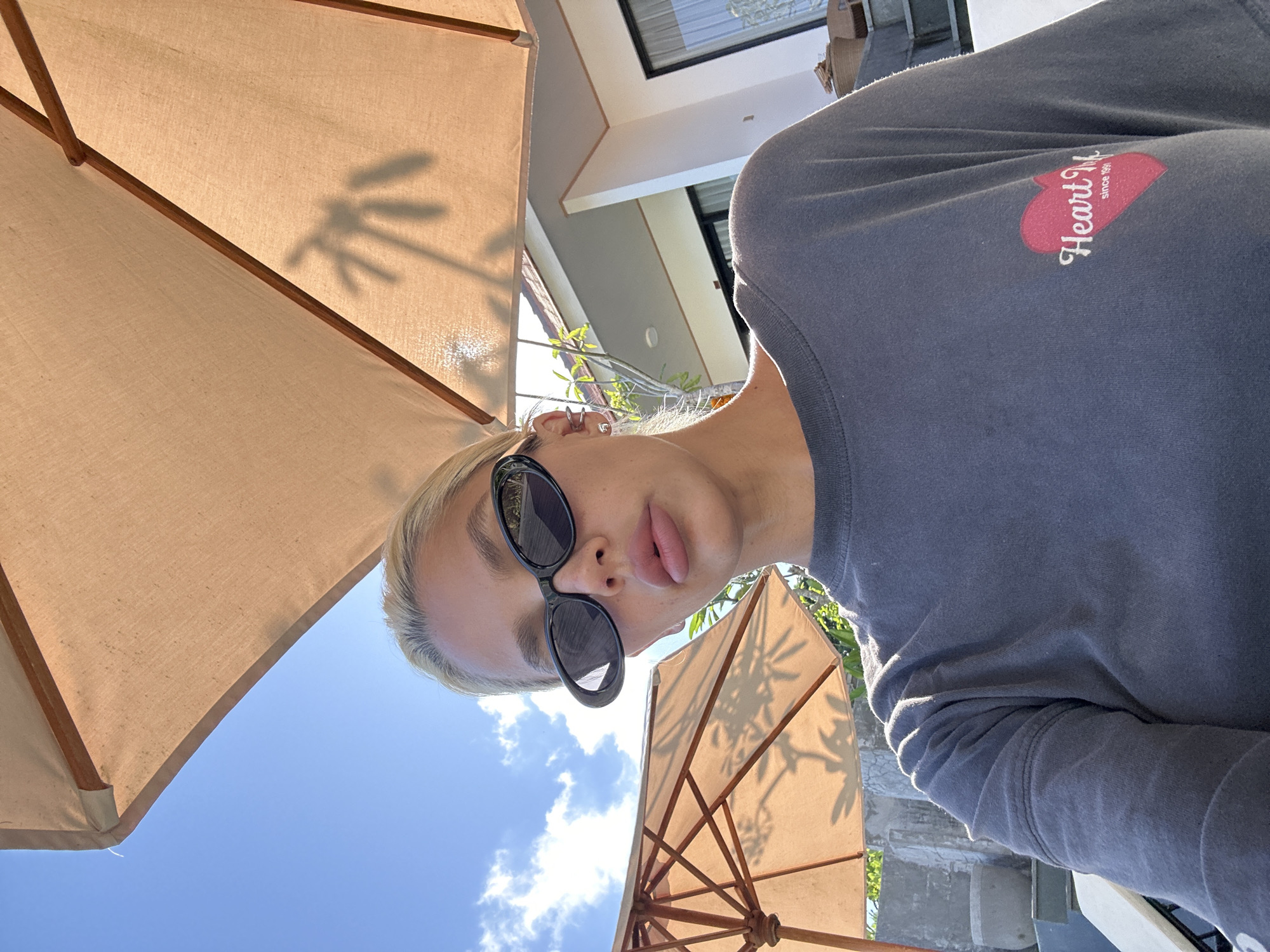 A close-up, low-angle selfie shows a young woman with blonde hair and sunglasses, wearing a dark grey t-shirt with a pink heart logo. She is outdoors, under a light brown umbrella, with a clear blue sky and scattered clouds in the background. The background also features a white building and lush green foliage, suggesting a tropical location. The lighting indicates it is daytime, likely sunny. The woman is looking towards the camera, with a neutral expression. The overall scene depicts a relaxed, casual moment in what appears to be a resort or outdoor leisure area. The specific location is in the city of Kuta Selatan, Indonesia.