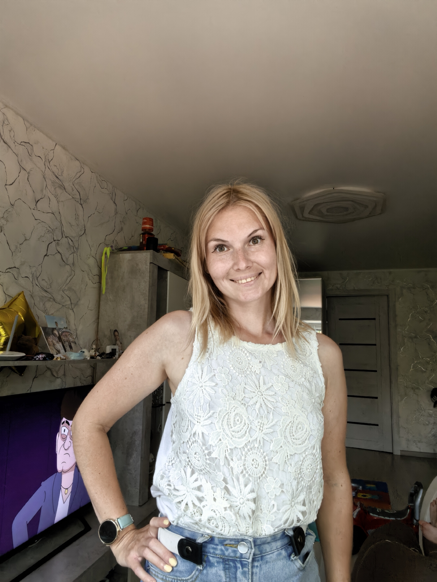 A woman with blonde hair and freckles smiles at the camera, wearing a white lace top and denim shorts. She is standing in a room with textured walls and a hexagonal ceiling fixture. To her left, a television screen displays a cartoon character. A grey cabinet and a stack of items are visible in the background. The scene appears to be an indoor residential setting.