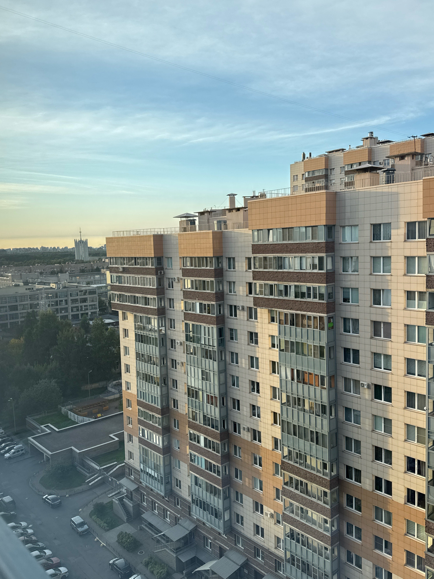 An elevated, eye-level, vertical shot captures a modern apartment building bathed in the warm glow of late afternoon sun. The building's facade is a grid of light beige and brown tiles interspersed with rows of windows, some reflecting the sky and others revealing interior lights. A prominent glass-enclosed section runs vertically along one corner, showcasing multiple balconies and the building's interior. The sky above is a soft blue with wispy white clouds and two thin, parallel power lines stretching across it. In the distance, other buildings and a tall, spire-like structure are visible, suggesting an urban environment. The lower portion of the image reveals a street with parked cars and a playground area adjacent to the building. The scene is calm and devoid of human presence.