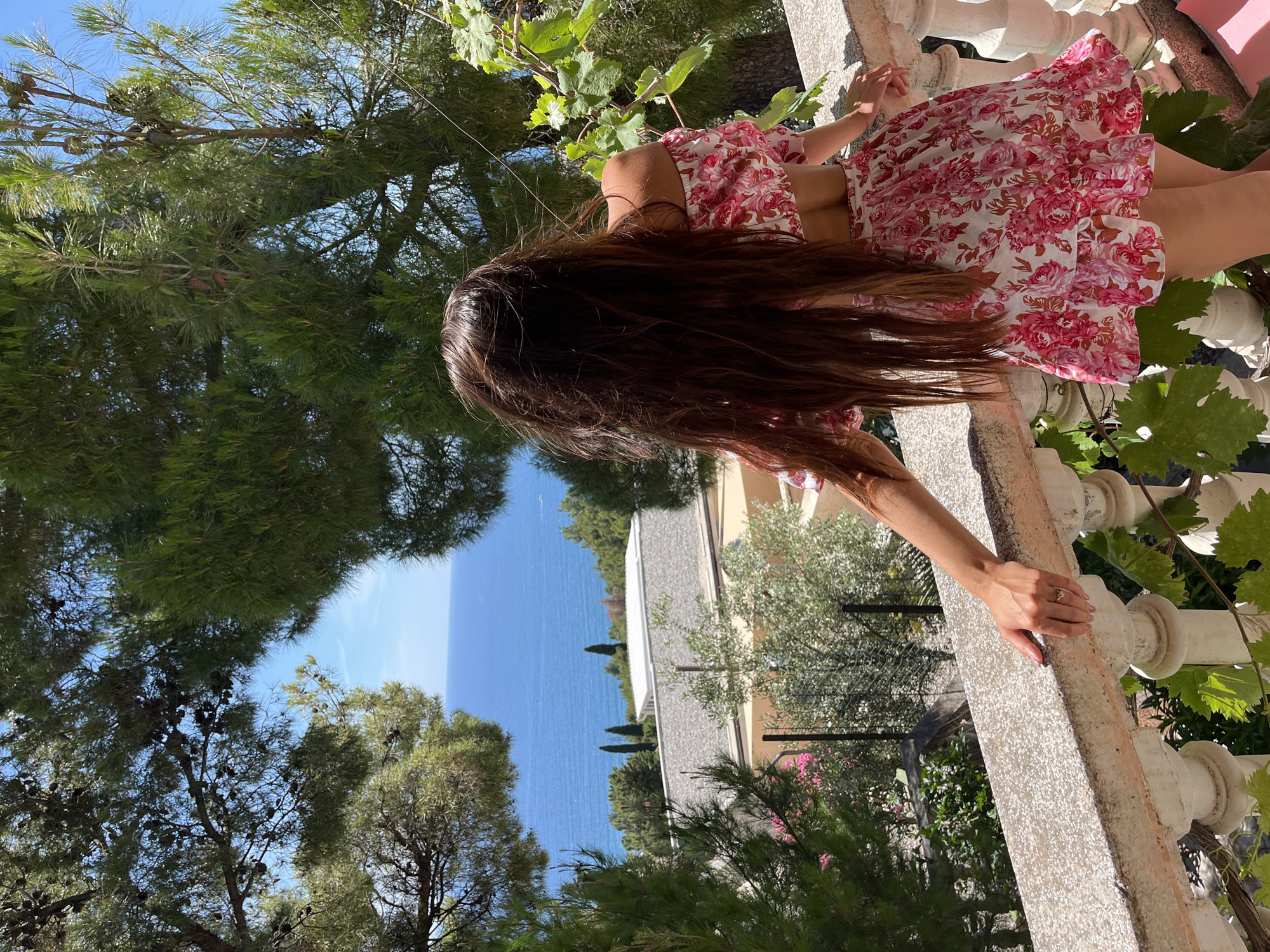 A young woman with long, dark brown hair stands on a balcony overlooking the Adriatic Sea in Šušanj, Montenegro. She is wearing a pink floral off-the-shoulder crop top and a matching skirt. The balcony has a white stone railing and is surrounded by lush green trees and vines. In the distance, buildings and a few cypress trees are visible against the blue water and sky. The woman is leaning on the railing, facing away from the camera, her hair gently blowing in the breeze.