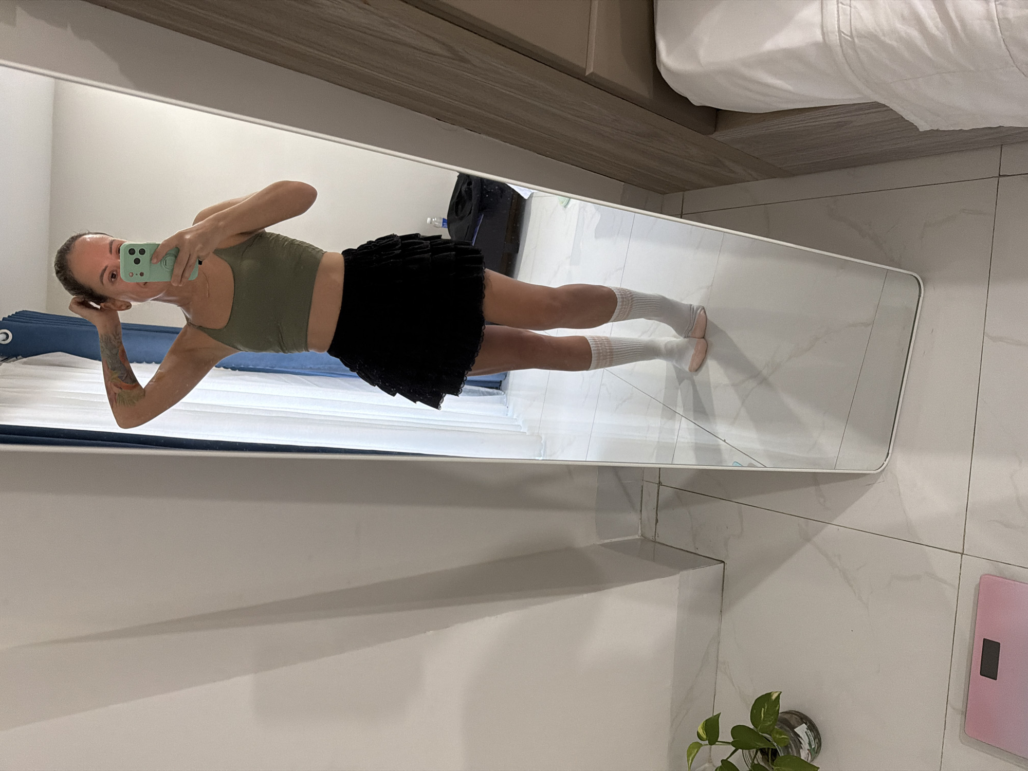 The image shows a woman taking a mirror selfie in a bedroom. She is wearing a green tank top, a black pleated skirt, and white socks. Her left arm is resting on her head, and she is holding a light green smartphone in her right hand, which is angled to capture her reflection. The room has white tiled floors and a large mirror that extends from the floor to near the ceiling. In the reflection, a dark suitcase and a portion of a white bed are visible. The lighting suggests it could be daytime. A pink scale and a potted plant are on the floor to the right of the mirror. The scene appears to be a personal, casual moment.
