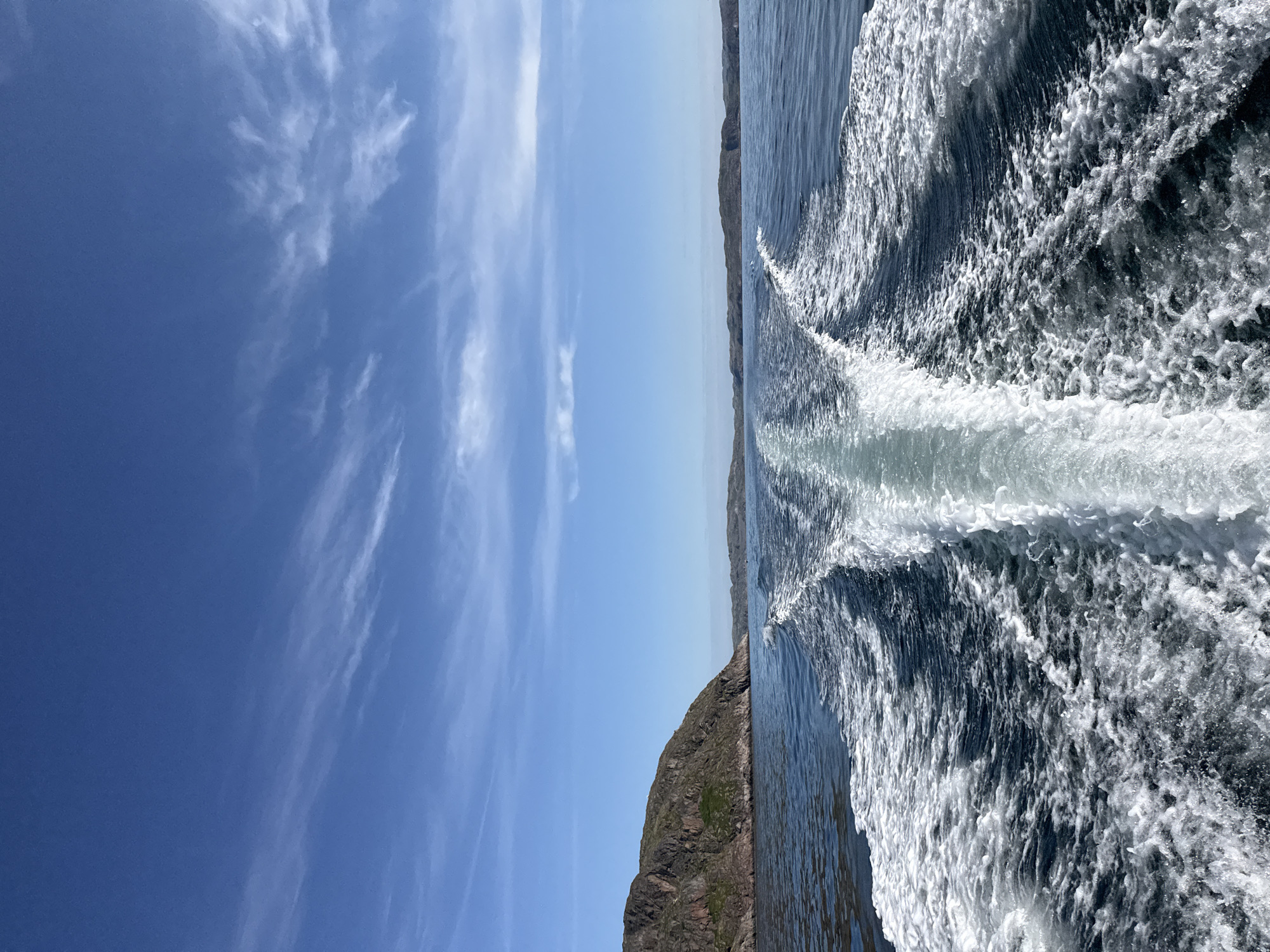 The image depicts a rear view from a moving boat on a body of water, likely the Kola Bay in Teriberka, Russia, given the terrain. The water is dark blue and disturbed, with a prominent white wake trailing behind the vessel. The wake fans out and churns with white foam. On the left side of the frame, a rocky, sparsely vegetated hillside rises from the water, characteristic of the Arctic coastal landscape. The sky is a clear, bright blue with scattered, wispy white clouds. In the distance, the shoreline is visible as a series of low, undulating hills or islands. The scene conveys a sense of travel and exploration in a rugged, natural environment.