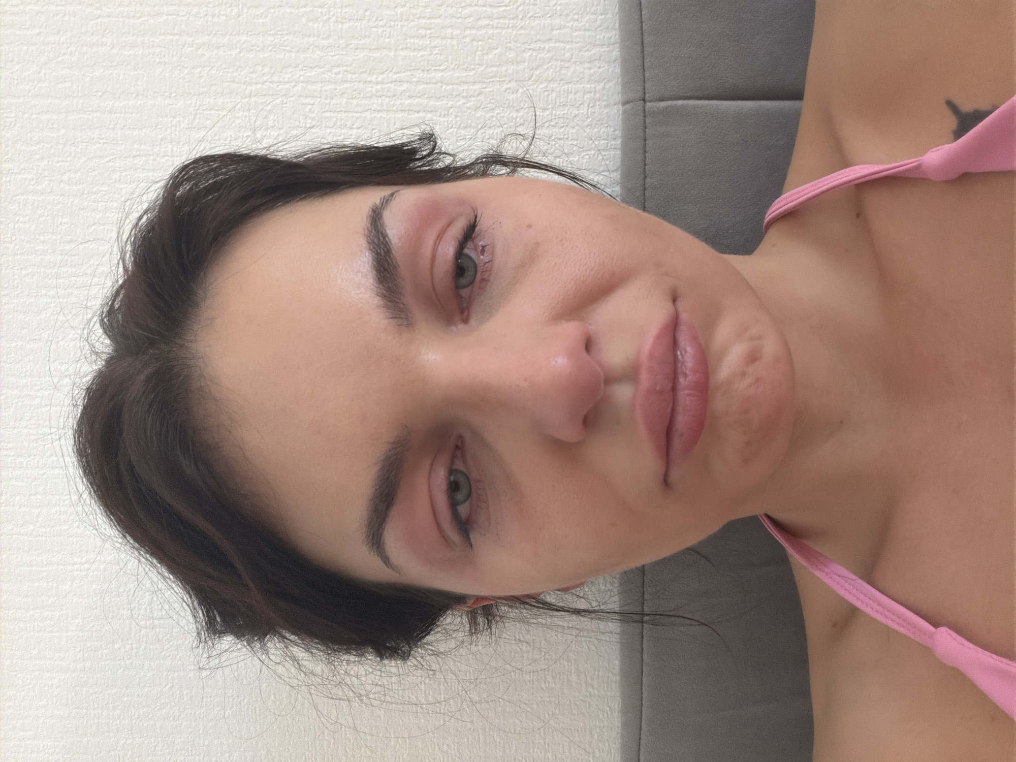 A close-up, front-facing shot features a woman with dark hair, her face partially obscured by the top of a gray headboard. She is wearing a pink spaghetti strap top. Her eyes are red and appear swollen, with pinkish discoloration around them. Her lips are slightly pursed. The background is a textured, off-white wall. The scene is indoors and does not provide any information about a specific location in Saint Petersburg, Russia.