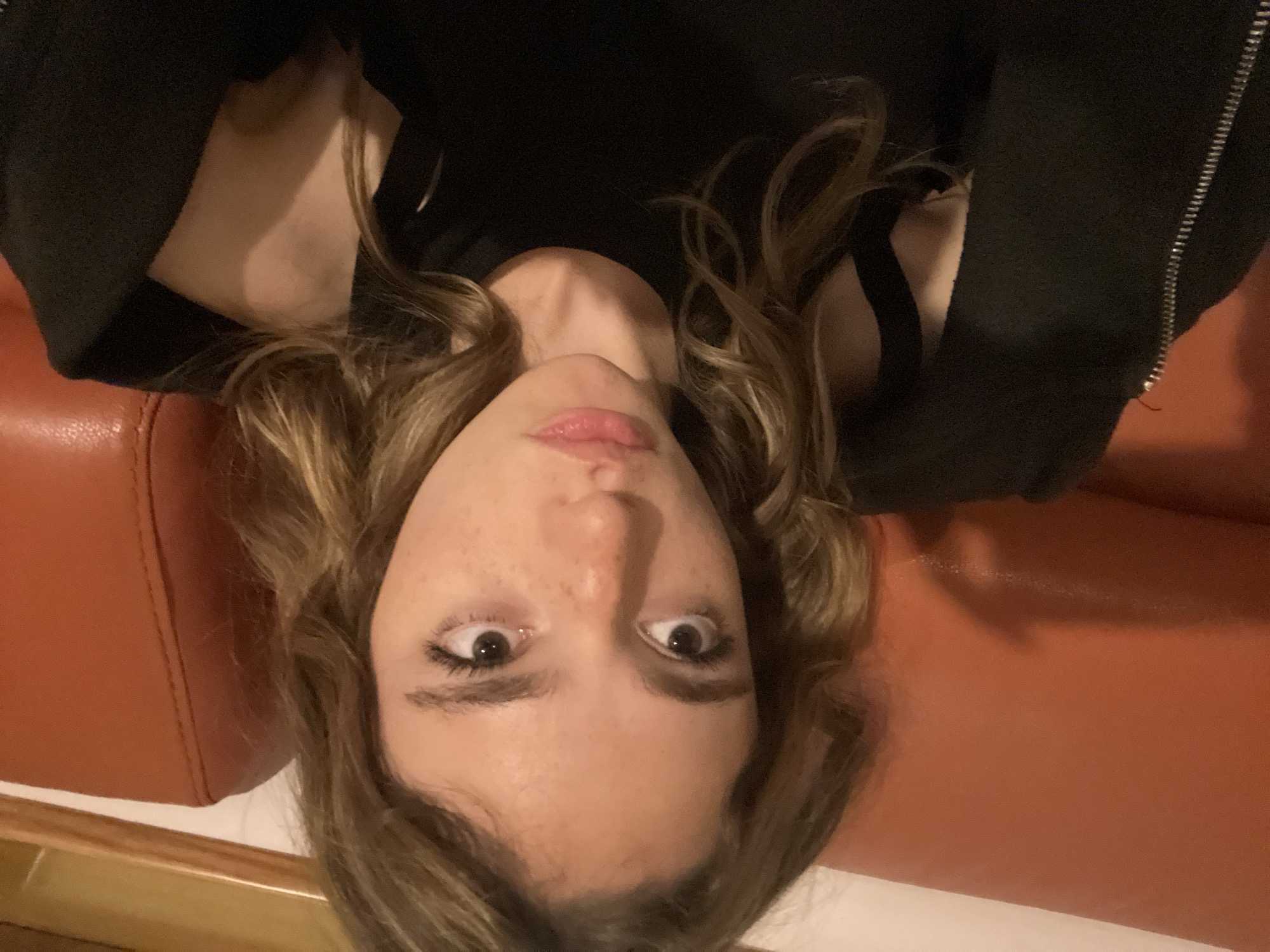A young woman with light brown, curly hair is lying upside down on what appears to be a brown leather couch or surface. She is wearing a black garment with shoulder straps visible. The environment around her includes a white surface below the couch and a glimpse of wood trim. There are no immediate indicators pinpointing the location as San Bernardino Tlaxcalancingo, Mexico.