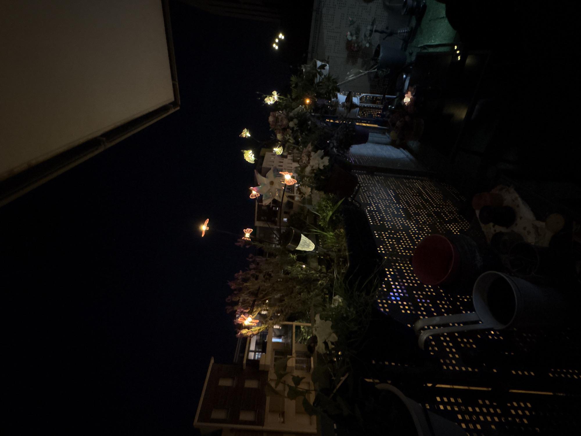 The image shows an outdoor setting in Strasbourg, France, at night. The foreground features a perforated metal table with various items, including a red bucket and a grey container, possibly a watering can. Decorative floral arrangements with small, glowing butterflies are present, adding to the ambiance. In the background, apartment buildings are partially visible, illuminated by interior lights. The sky is dark, with a few distant lights. The scene suggests a balcony or rooftop garden with urban surroundings.