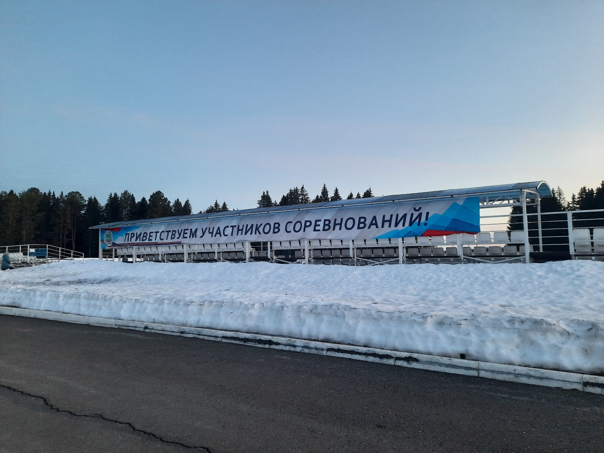 The image shows a winter scene in what appears to be the city of Krasnogorskoye, Russia. A snow-covered grandstand is prominent in the midground, with a large banner reading "ПРИВЕТСТВУЕМ УЧАСТНИКОВ СОРЕВНОВАНИЙ!" (Welcome Participants of Competitions!) across its front. Behind the grandstand are evergreen trees, suggesting a forested or park area. In the foreground is an asphalt road. The sky is a gradient of light blue to a softer, pale shade near the horizon, indicating dawn or dusk. There are no people visible in the frame. The environment is consistent with a cold climate and winter conditions.
