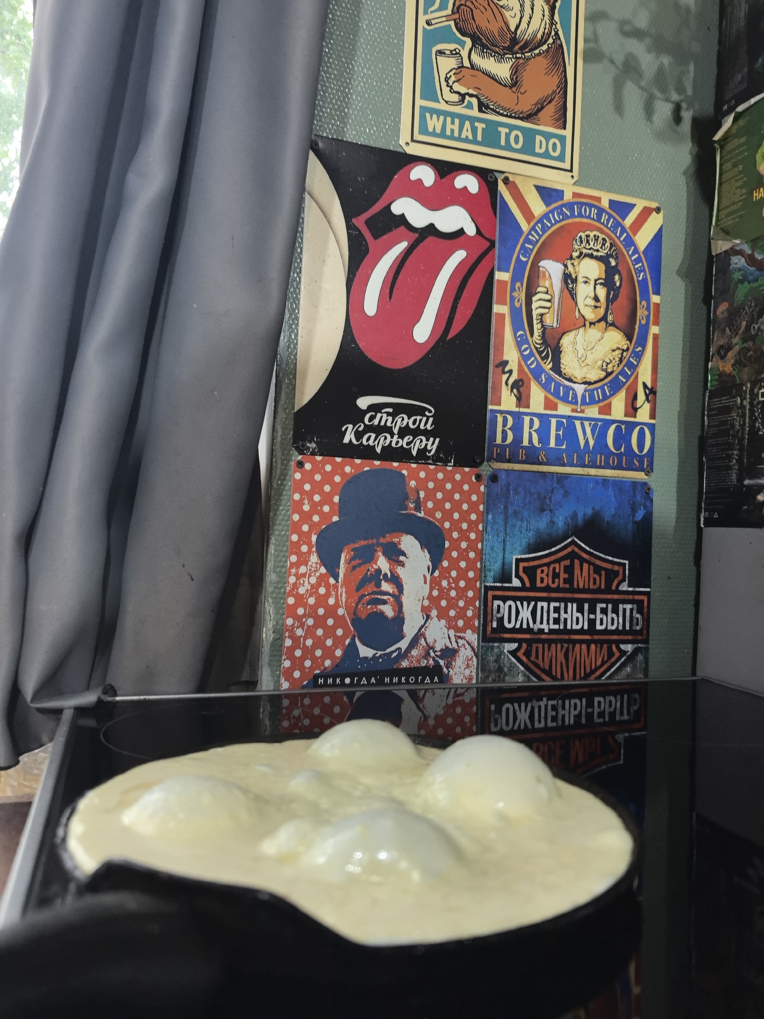A close-up shows a black pan on a stovetop containing what appears to be cooking eggs. Behind the stovetop, a wall is decorated with various posters, including a Rolling Stones tongue logo, Queen Elizabeth holding a beer, and an image of Winston Churchill. The text on other posters is in Russian. To the left, a grey curtain hangs. The environment suggests a kitchen setting. The city of Noviy, Russia is not apparent.