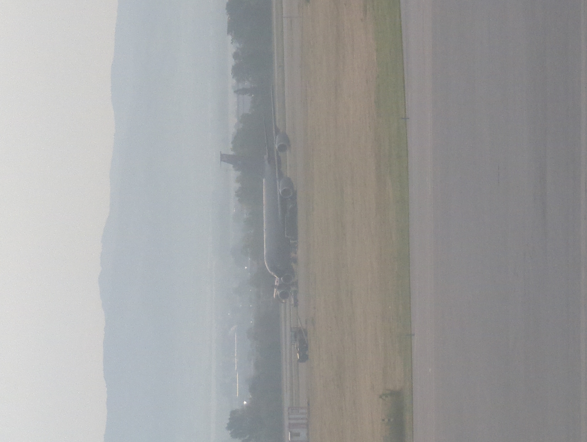 The image captures a hazy, overcast scene at an airport. In the foreground, a paved surface, likely a taxiway or runway, stretches across the frame. Beyond this, a grassy field extends towards a larger aircraft parked on the tarmac. The aircraft is a wide-body jet with multiple engines visible under its wings. To the left of the aircraft, there are several structures, including what appears to be a small building or shelter, and a vehicle that resembles a tow truck. In the background, a line of trees partially obscures more distant buildings and a hazy mountain range is visible on the horizon. The overall atmosphere is muted due to the atmospheric conditions, suggesting low visibility. The location is identified as Sofia, Bulgaria.