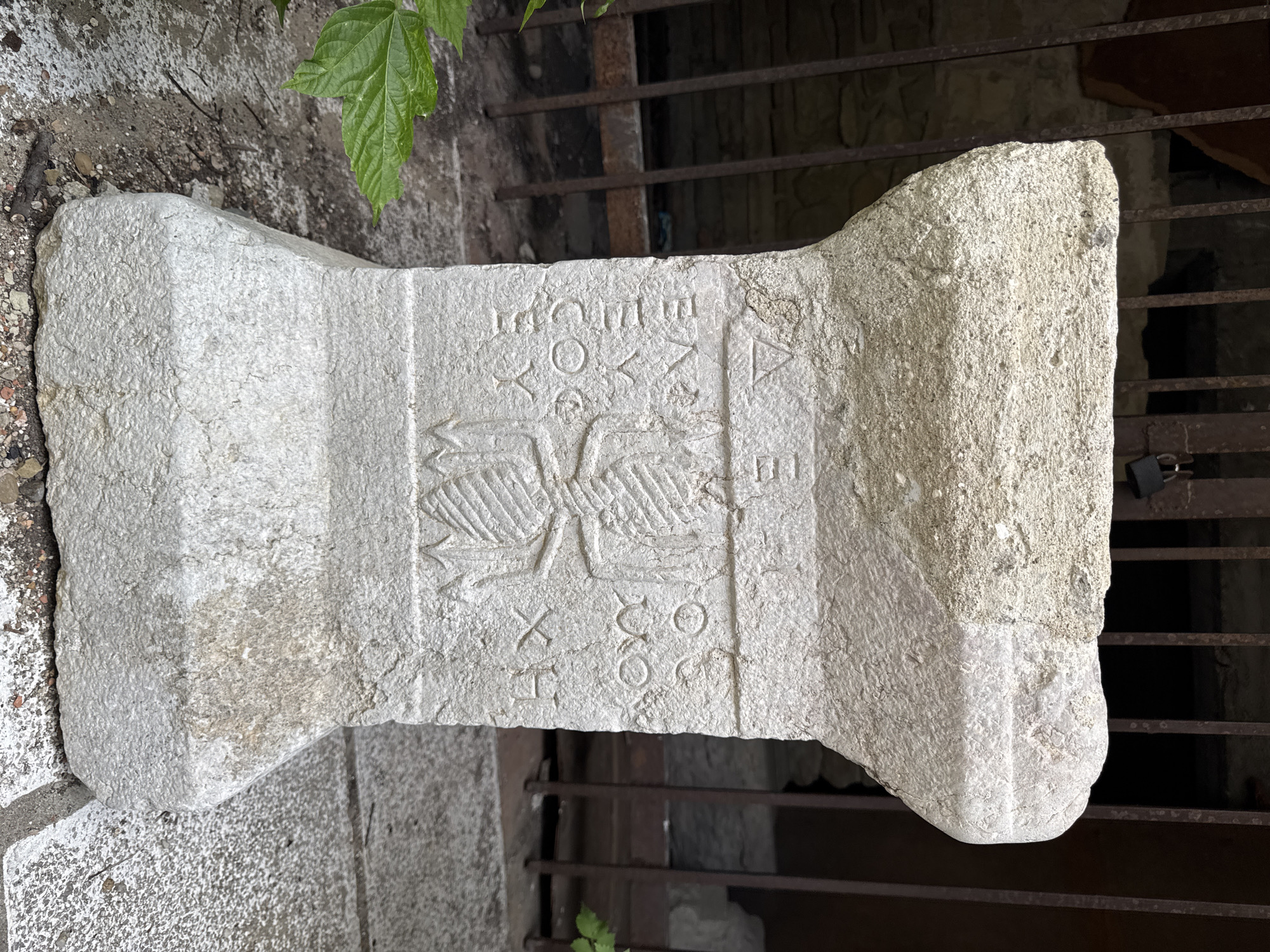 The image shows a close-up of an ancient stone artifact with inscriptions. The artifact is weathered and appears to be made of light-colored stone, possibly limestone. It has a roughly rectangular base and a wider, flat top. The central portion of the artifact features carved symbols and what appears to be a stylized insect or plant motif. Faint Greek or Old Bulgarian characters are inscribed on either side of this central image.
The artifact is situated outdoors, with a rusty metal grate or fence visible in the background. The ground around the artifact is covered with gravel and dirt. Green leaves from a plant are visible in the lower left corner, partially obscuring the artifact's base. The overall environment suggests an archaeological site or an outdoor display of historical objects.
This artifact is located in Veliko Tarnovo, Bulgaria, specifically within the Tsarevets fortress complex, which served as the capital of the Second Bulgarian Empire. It is likely a part of the historical remnants displayed within the fortress grounds.