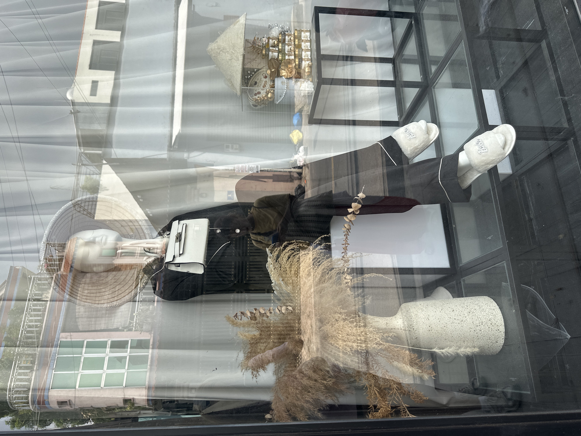 A shop window display in Đồng Nai, Vietnam features a mannequin wearing a conical hat and black pajamas with white slippers. The mannequin holds a small white purse. The display also includes a vase with dried grasses and a decorative gift set. The window reflects a building across the street.