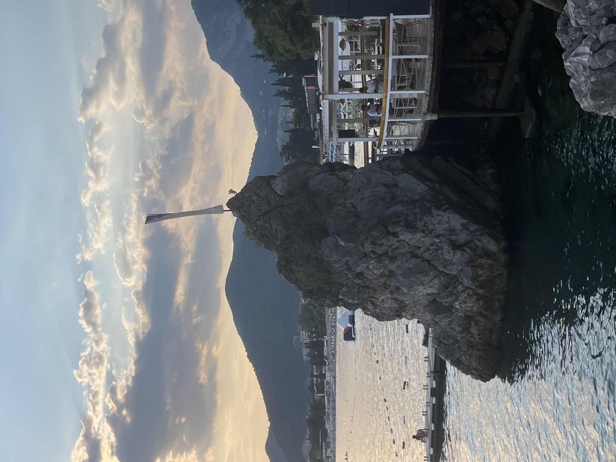 A scenic view of a coastal town with a restaurant built on stilts over the water, in Budva, Montenegro. The scene captures a rocky outcrop in the foreground, with a white flag planted on its summit. Behind the rock, a white, open-air restaurant with tables and chairs is visible, with people seated at some of them. The restaurant is elevated and appears to be overlooking a body of water. In the background, a beach with umbrellas and a few people can be seen, leading to a town with buildings nestled against a backdrop of rolling, green mountains. The sky is filled with dramatic clouds, suggesting it is either sunrise or sunset, casting a warm glow over the scene. The water is calm and reflects the sky.