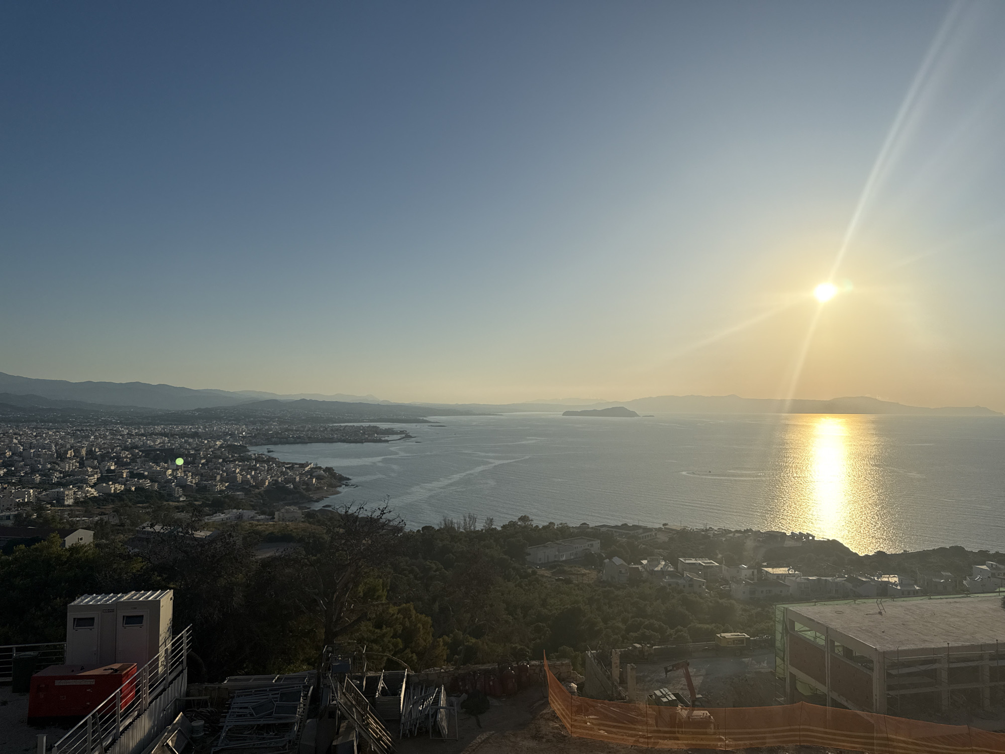 This image depicts a panoramic view of a coastal city at sunrise or sunset. The sun, a bright orb, is positioned in the upper right quadrant of the image, casting a golden reflection across the calm, rippling sea. In the foreground, a construction site is visible with scaffolding, orange safety netting, and building materials. Several portable toilet structures and a red generator unit are also present. Beyond the construction site, the city sprawls towards the coastline, characterized by densely packed buildings with white and light-colored roofs. A bay or harbor is present, with boats or ships potentially dotting the water. In the distance, rolling hills and mountains form the backdrop to the city and the sea, with a hazy atmosphere suggesting either pollution or the atmospheric effects of the low sun angle. The sky is a clear, gradient blue, transitioning to a pale yellow near the horizon. There are no people visible in the scene.