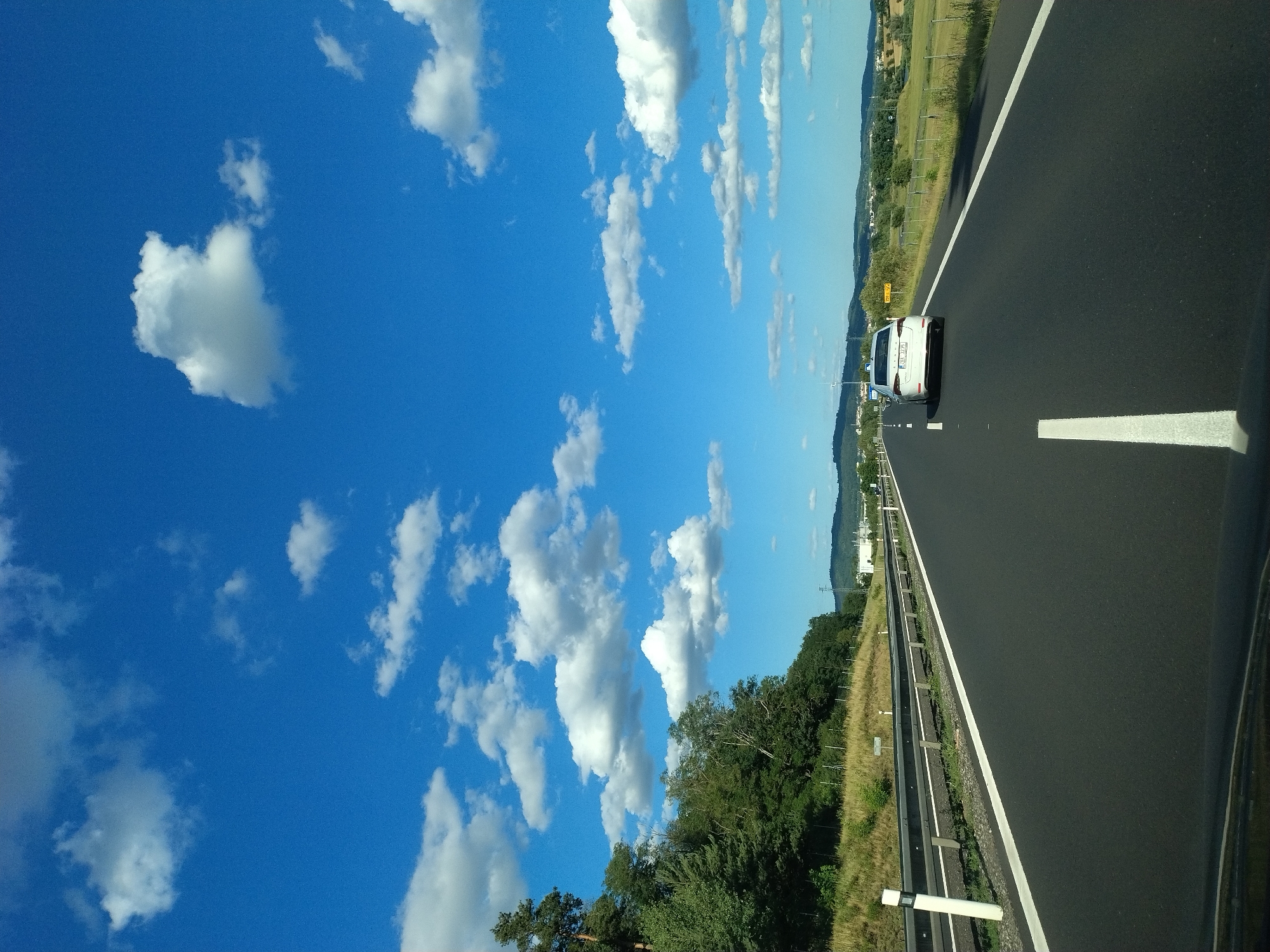 The image shows a view from the driver's seat of a vehicle traveling on a highway. The highway has a black asphalt surface with clear white lane markings. To the left of the highway, there is a guardrail and a grassy embankment with trees and vegetation. In the distance, there are hills and a small town or village visible. The sky is a clear blue with scattered white clouds. A white car is visible ahead in the right lane of the highway. A yellow sign with black text is also visible on the right side of the highway. The image is taken during daylight hours. The location is Krašovice, Czechia.