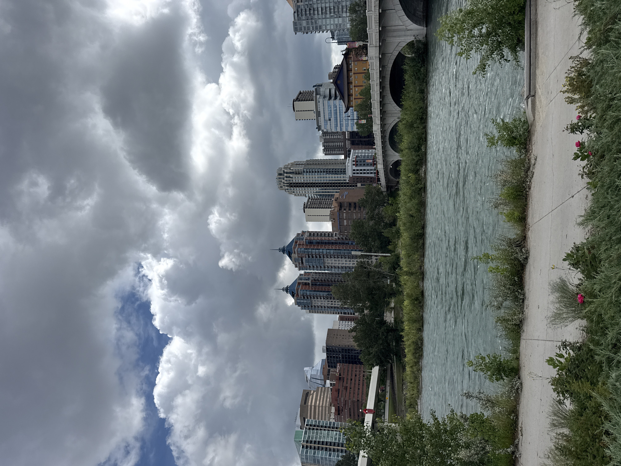 The image captures a view of the Calgary skyline from across the Bow River. In the foreground, a paved walkway borders the river, with lush green vegetation and a few scattered pink flowers. The Bow River itself is wide and appears to be flowing with moderate speed, its surface showing ripples and slight choppiness under a cloudy sky.
Dominating the mid-ground is a large, arched stone bridge spanning the river. Beyond the bridge, the Calgary skyline is visible, featuring a collection of modern high-rise buildings of varying architectural styles and heights. Among the prominent structures are several residential towers with distinct blue-capped roofs, as well as office buildings with glass facades. The sky above is filled with dramatic, dark gray clouds interspersed with patches of brighter sky, suggesting an overcast day with potential for rain. The overall scene presents a typical urban landscape in Calgary, Canada, with the river and bridge acting as natural separators between the park-like foreground and the bustling city center.