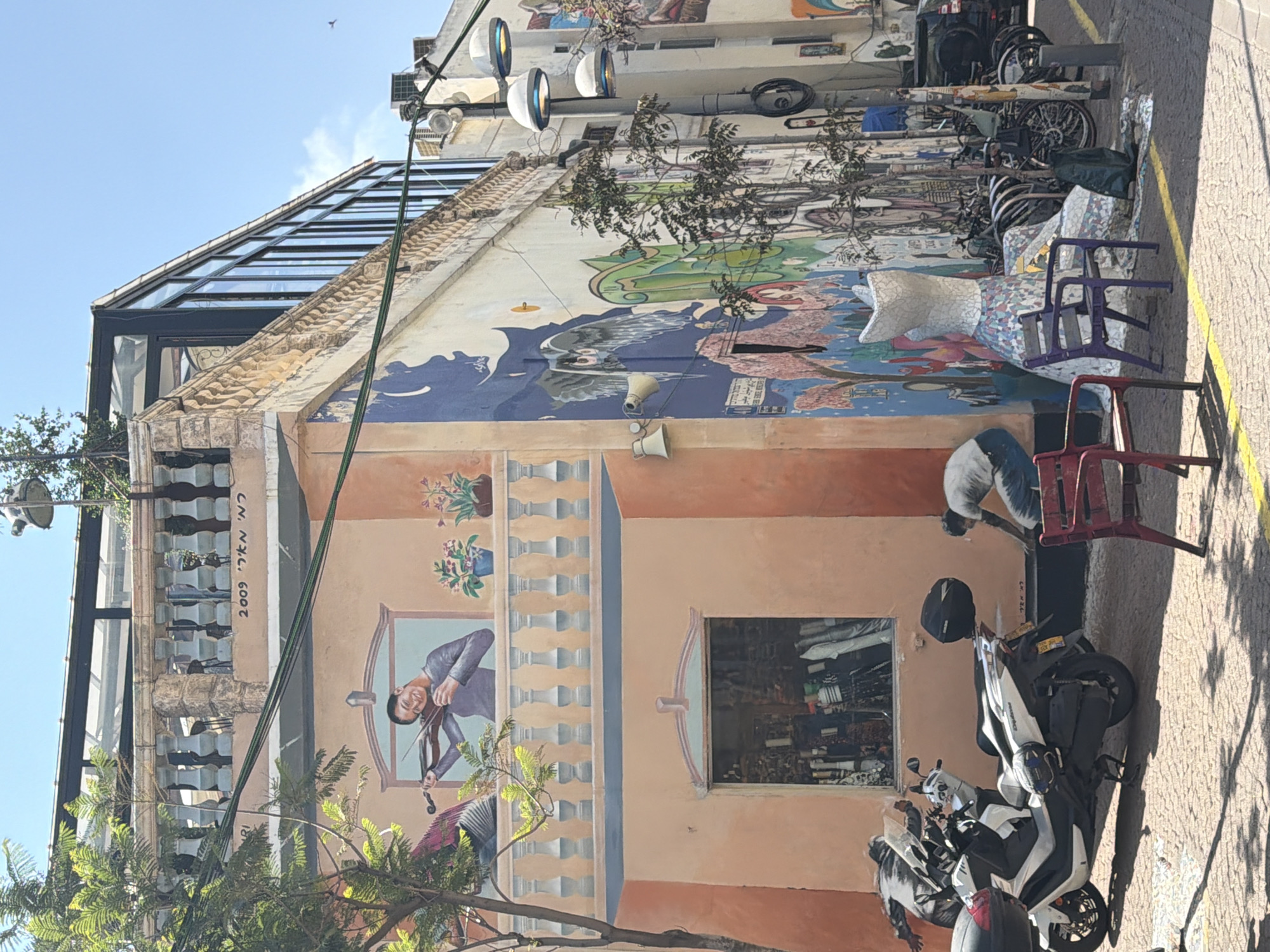 The image depicts a building facade in Tel Aviv, Israel, notable for its large mural. The mural features an orange and peach-colored section at the bottom, depicting a man playing a violin within a faux balcony. Above this, the mural transitions to a more abstract and colorful design featuring blues, whites, and earth tones.
A person is partially visible, leaning over near a red plastic chair, possibly interacting with something on the ground. Two motorcycles are parked in front of the building, a black helmet resting on one of them. Several bikes and objects are situated on the sidewalk next to the building.
Architecturally, the building has a mix of old and new elements. The mural is on a relatively older part of the building, while a modern glass and steel structure is visible on top.