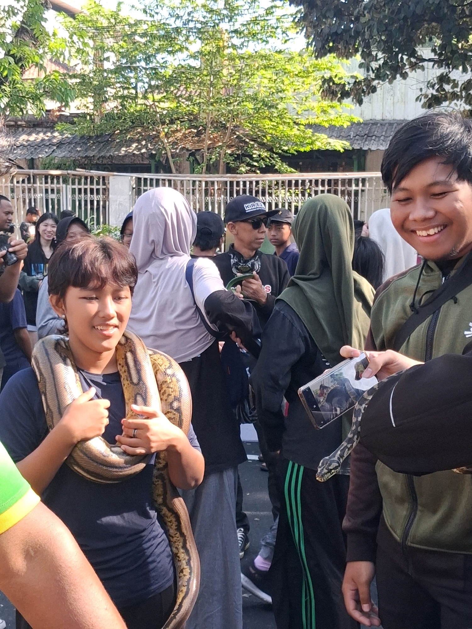 In the city of Surakarta, Indonesia, an outdoor gathering is depicted under a clear sky with sunlight filtering through lush green trees. The scene features a group of individuals, with a young person prominently positioned in the foreground, wearing a dark t-shirt and a large snake draped around their neck and shoulders. The reptile has a patterned skin in shades of brown and yellow. The individual holding the snake smiles and looks slightly to their left.
To the right of this central figure, another young person, wearing a dark green jacket, smiles broadly at the camera while holding a smartphone that displays an image of a snake. The phone is angled towards the snake around the other person's neck, suggesting an interaction of showing or documenting the animal.
Other people are visible in the background, some wearing headscarves and casual clothing. A white fence with a metal railing runs horizontally across the middle ground, separating the foreground action from buildings and foliage behind. The overall atmosphere appears to be casual and perhaps a local event or street scene.