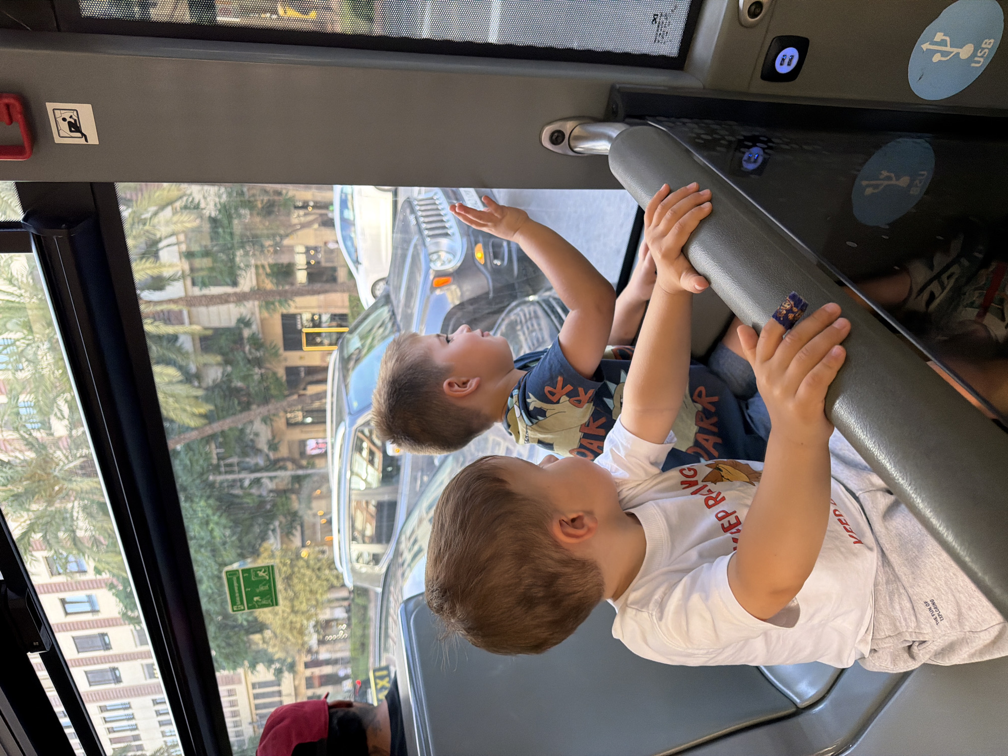 Two young boys are seated inside a bus, looking out the window. The boy in the foreground, on the left, is wearing a white t-shirt and light gray shorts. He is looking to his right. The boy in the background, on the right, is wearing a blue t-shirt with a graphic print. He is looking out the window and has his right hand raised. Outside the bus, palm trees and buildings are visible, suggesting an urban environment. The interior of the bus shows gray seating and a window with various stickers and signs on the wall. One sticker on the wall depicts a silhouette of a person, possibly indicating a safety instruction or a depiction of a passenger. Another green sticker displays icons that might relate to transit rules. USB charging ports are visible on the bus's interior paneling. The scene appears to be captured during daylight hours.