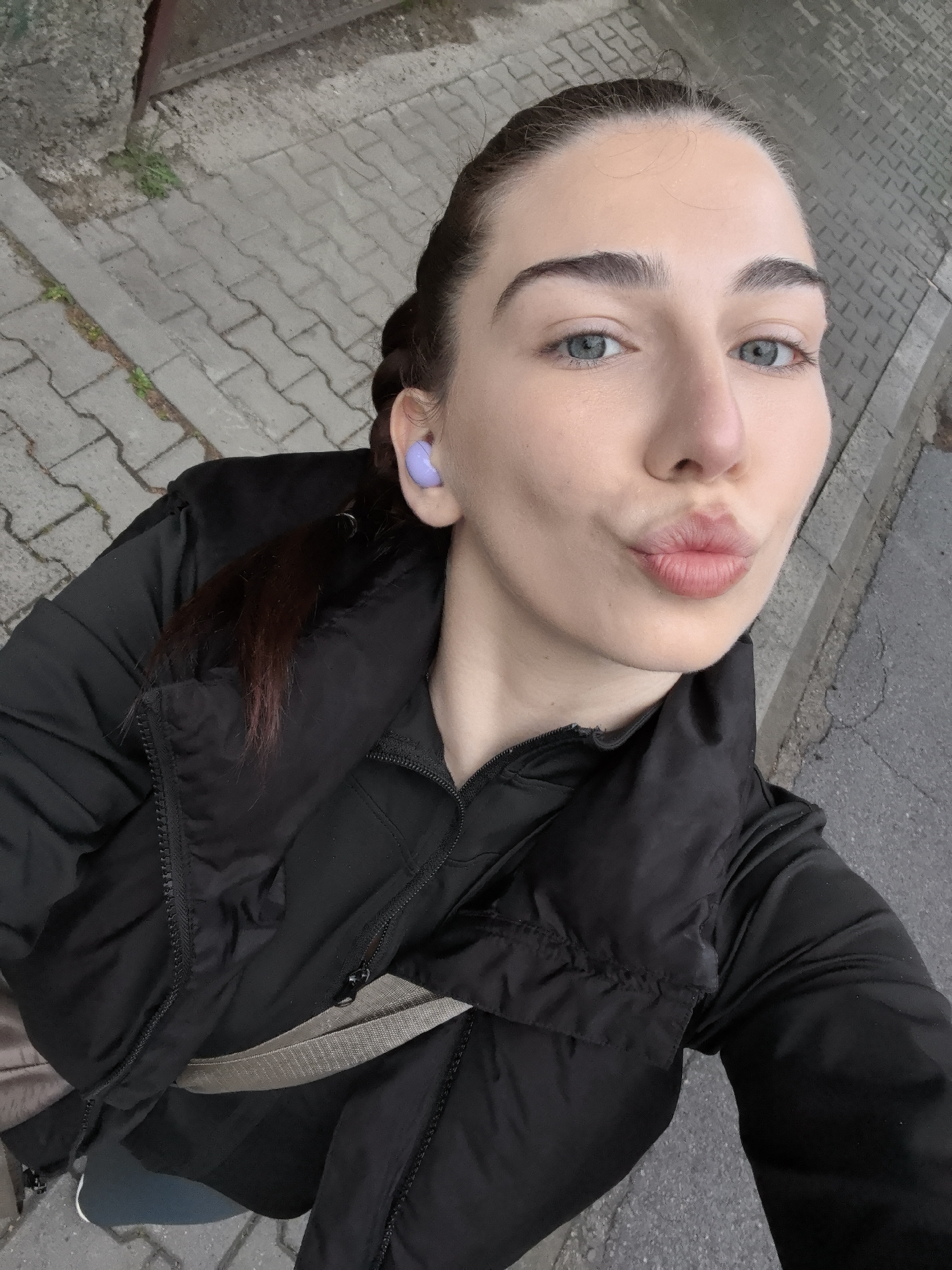 A close-up selfie captures a young woman outdoors, looking directly at the camera with a playful expression, her lips puckered into a 'kissy' face. She has light skin, prominent dark eyebrows, and striking blue-grey eyes. Her dark hair is styled in a braid, visible over her left shoulder. A bright purple earbud is visible in her right ear. She is wearing a black puffy jacket over a black long-sleeved top, with a light-colored strap, possibly from a bag, crossing her chest.
The background shows a paved sidewalk made of rectangular grey blocks, with sparse patches of green vegetation growing in cracks. Adjacent to the sidewalk, a darker asphalt surface or road is visible. A grey concrete wall or structure is also present in the upper left. The lighting is soft and even, suggesting an overcast day. No visible text is present in the image.